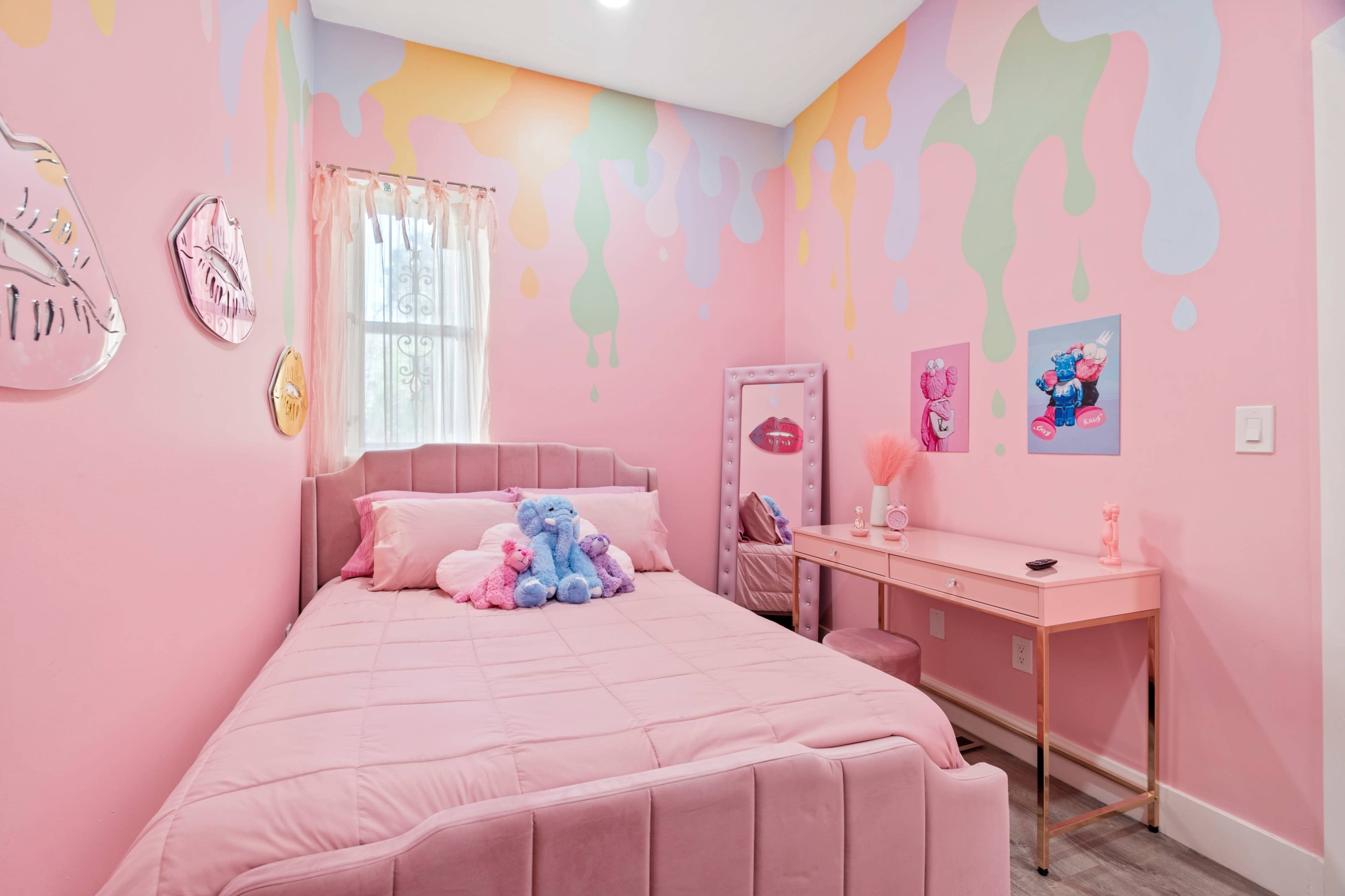 A small bedroom with pink walls featuring colorful drip patterns, a single bed adorned with plush toys, and a matching desk set with a mirror.