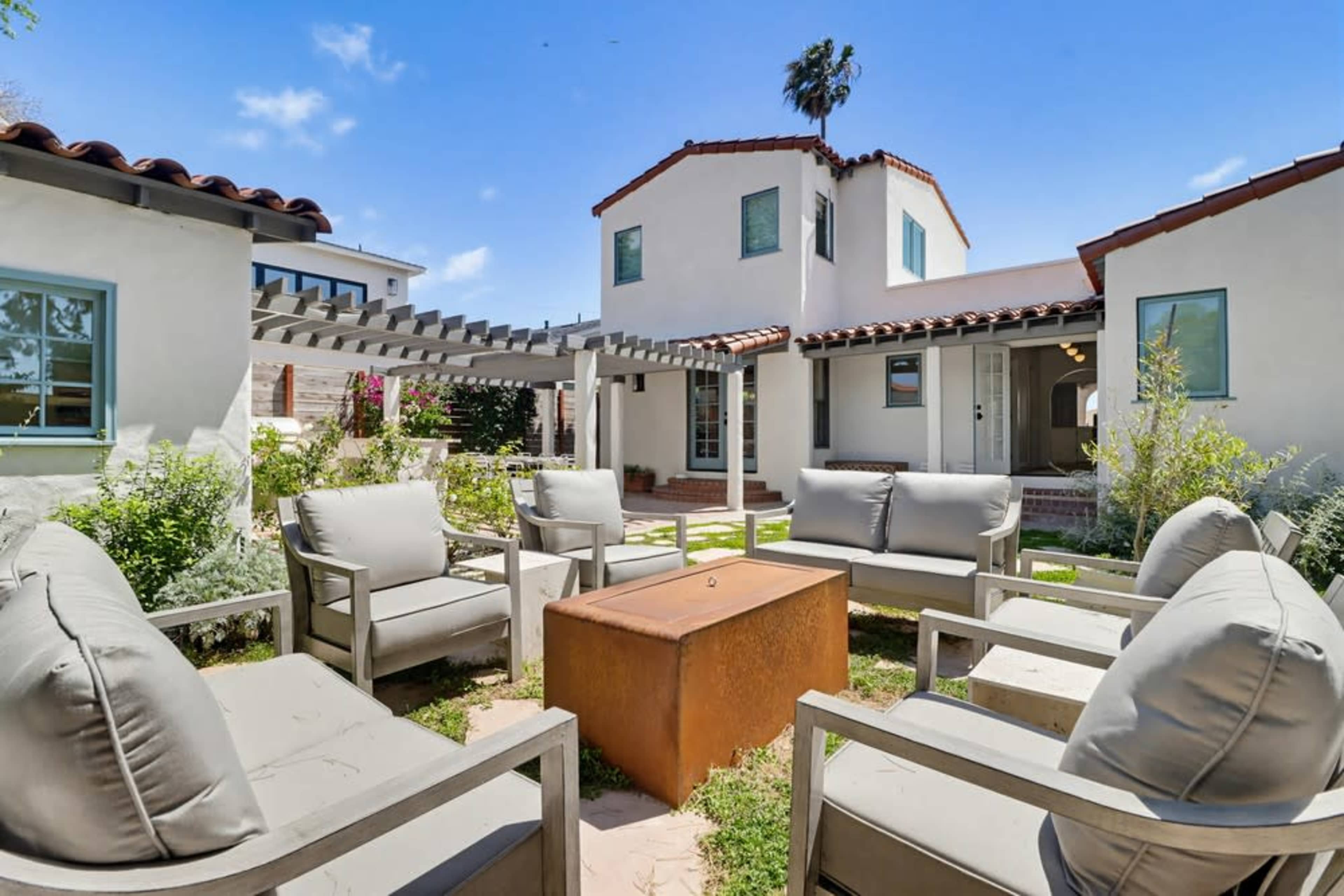 A courtyard features several gray lounge chairs arranged around a central fire pit, with a two-story building and lush greenery in the background.