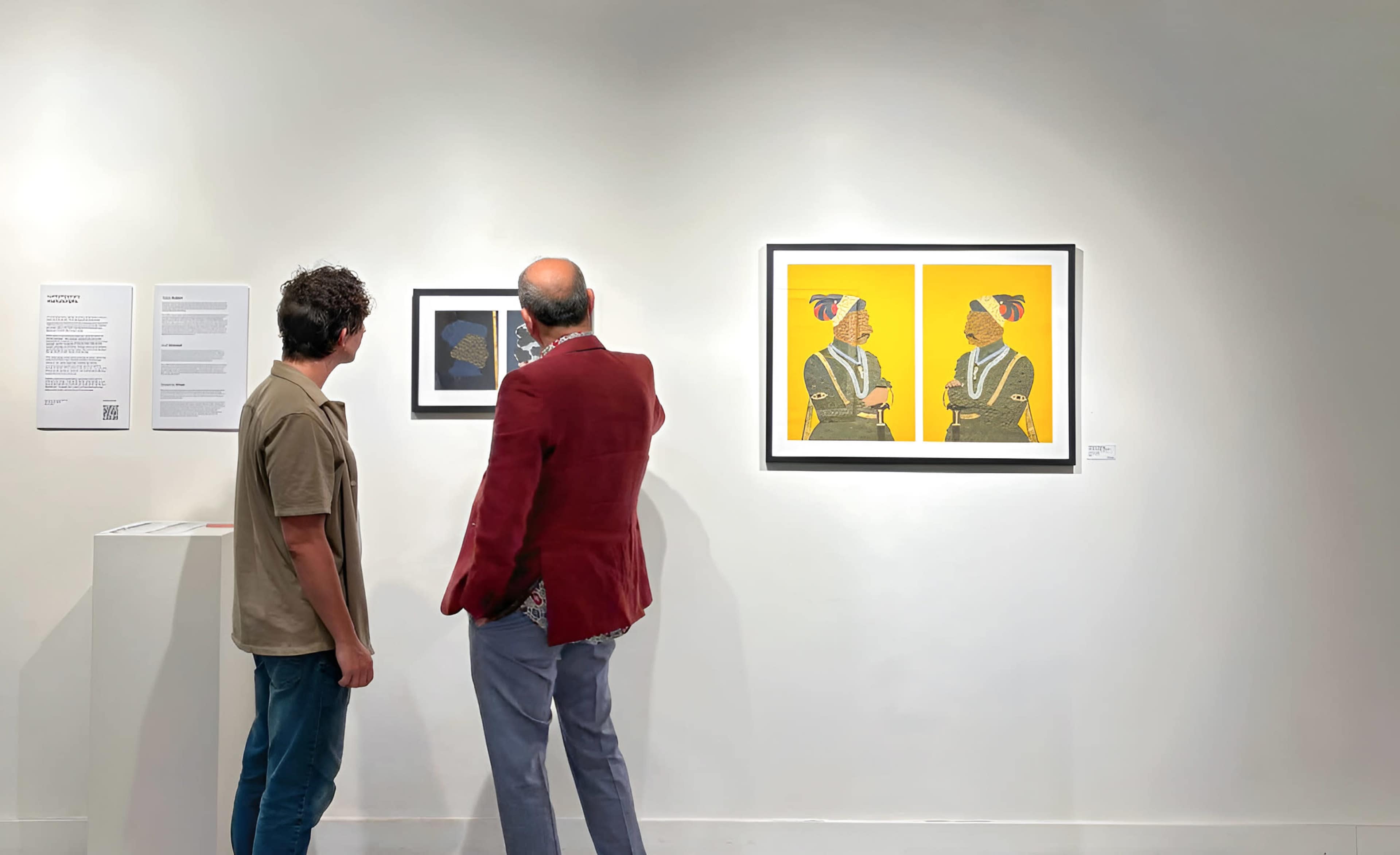 Two individuals are observing framed artworks on a white gallery wall, with informational text nearby.