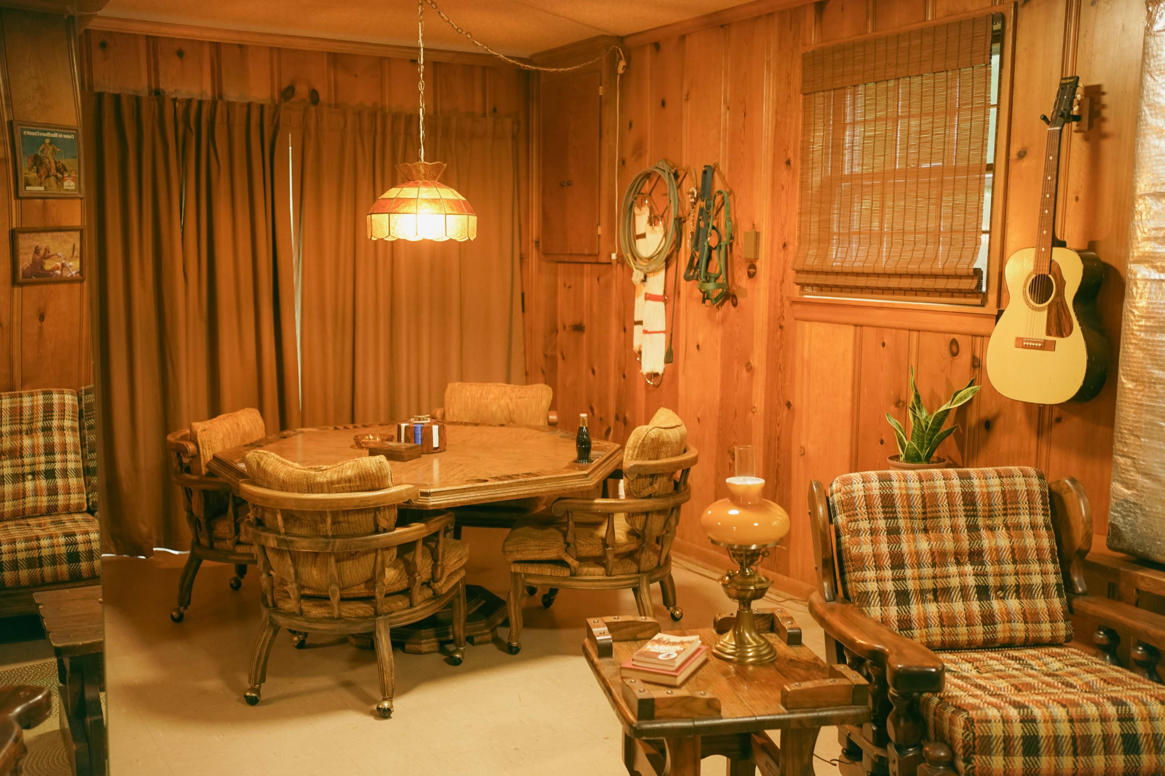 A wooden-paneled room features a round table surrounded by upholstered chairs, with a hanging light fixture and a guitar on the wall.