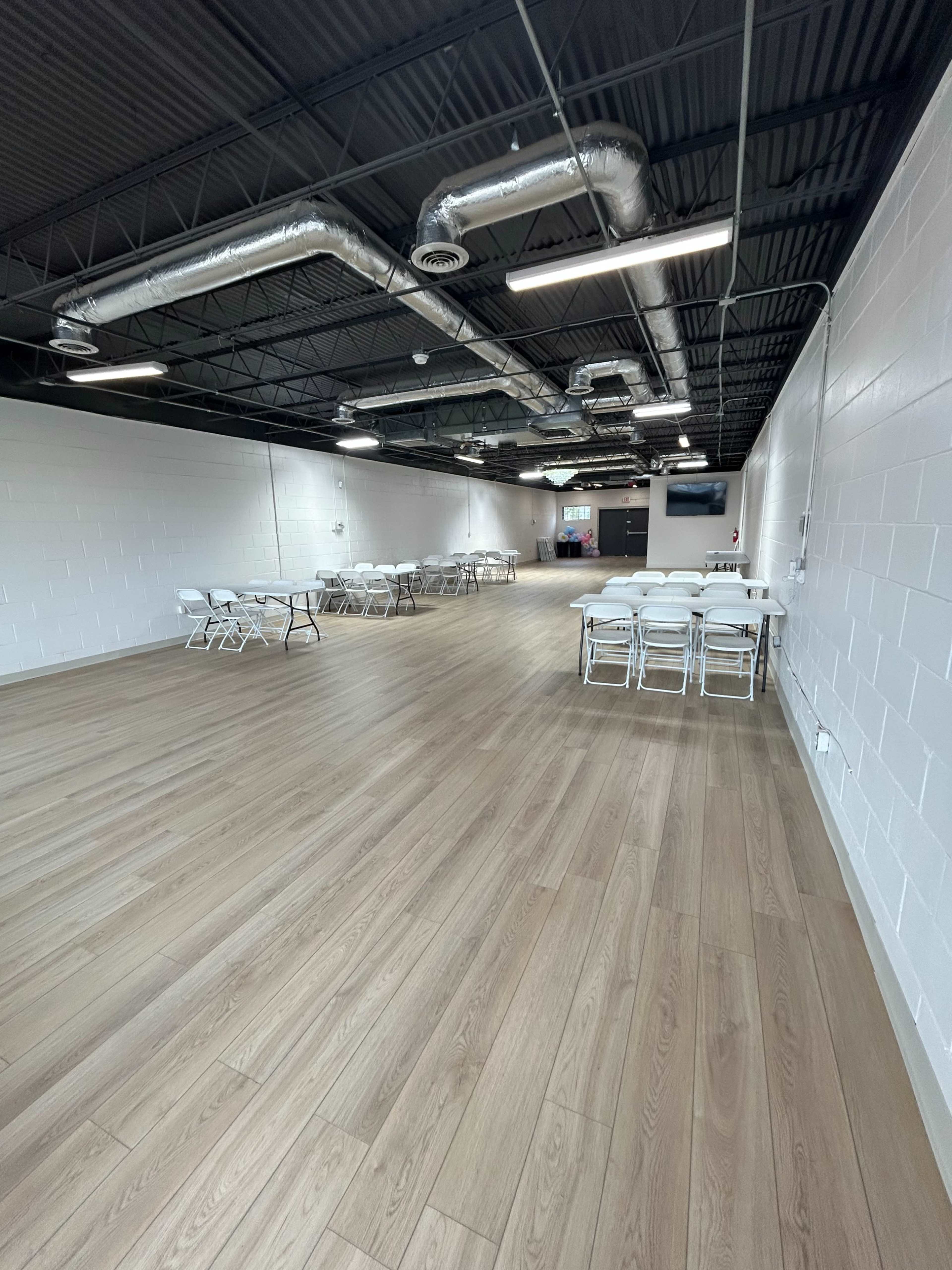 The image depicts a spacious, well-lit indoor area featuring rows of folding tables and chairs set up on a wooden floor.