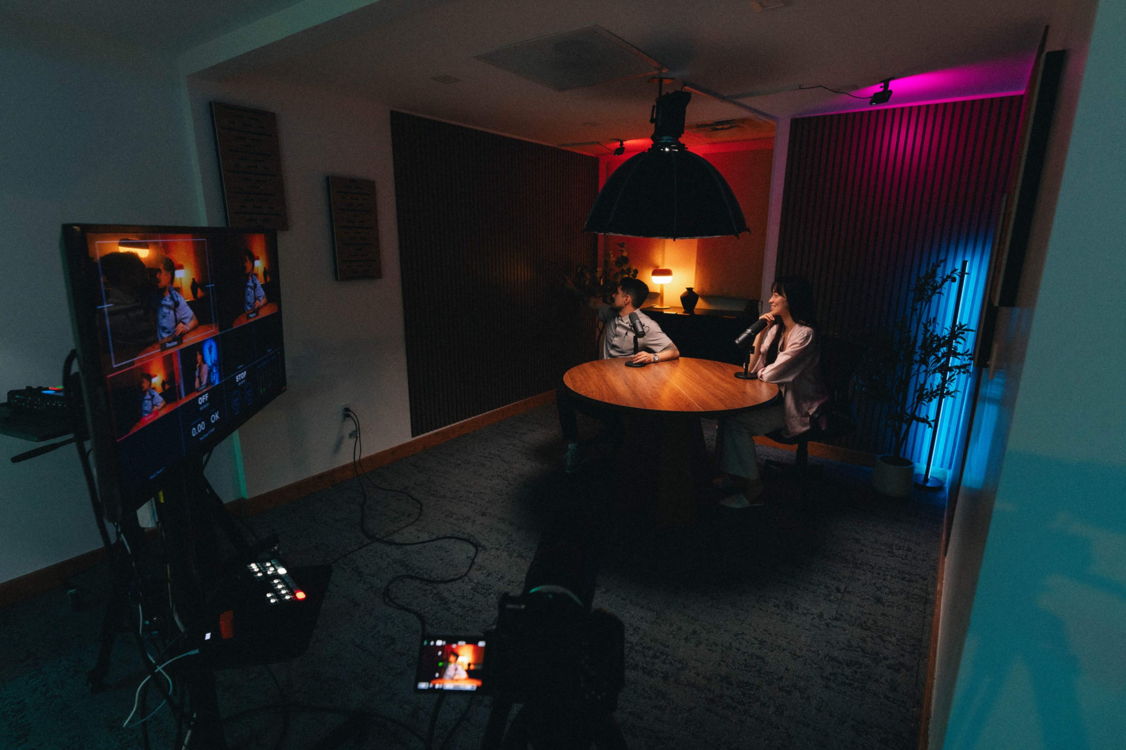 A round wooden table is set in a dimly lit room with two people seated across from each other, while a video camera and monitor display their conversation.