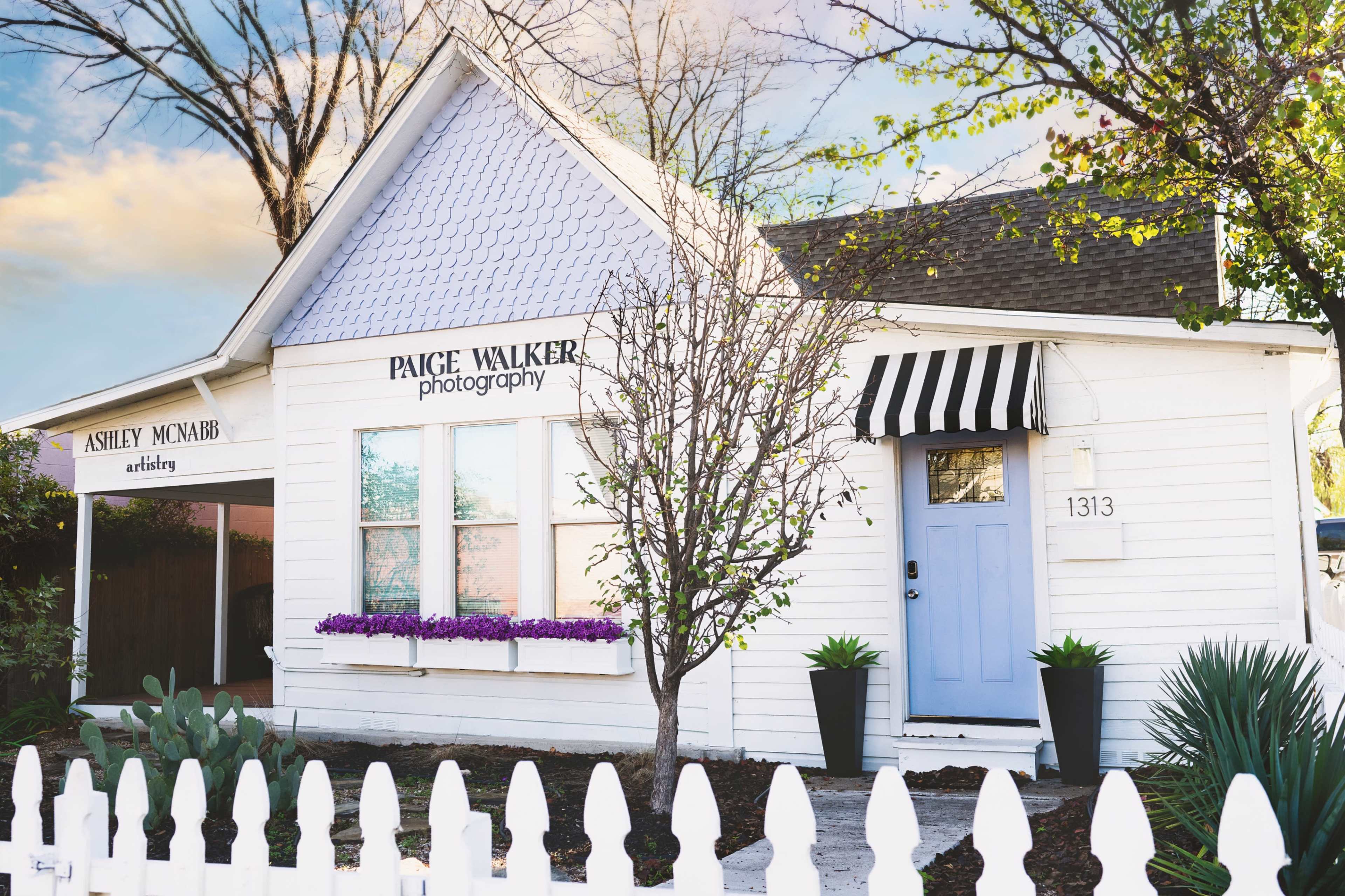 A white cottage with lavender accents and a striped awning houses Paige Walker Photography, alongside a separate space for Ashley McNairb Artistry.
