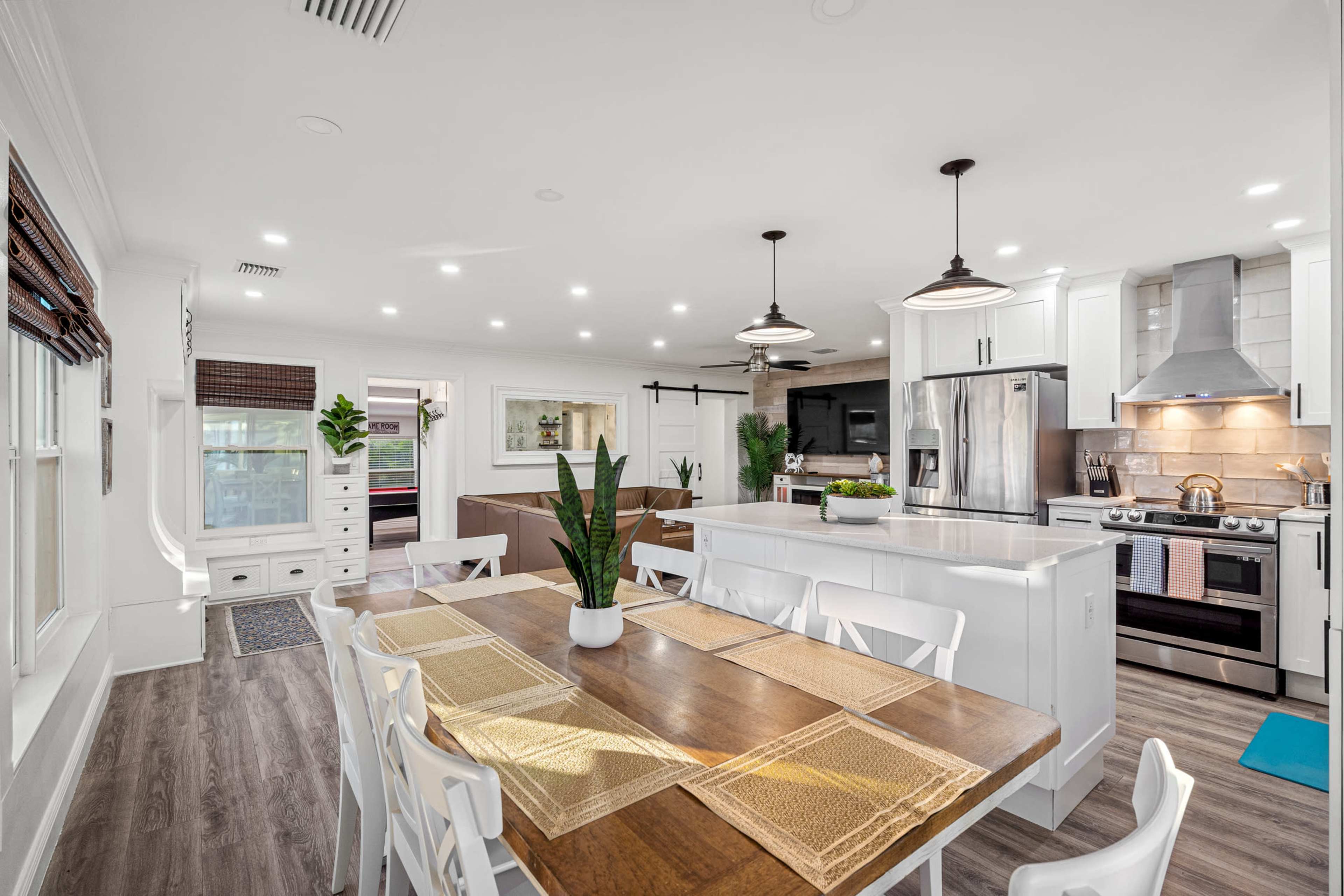 A bright, modern kitchen features a central island with a dining table and white cabinetry, illuminated by pendant lights.