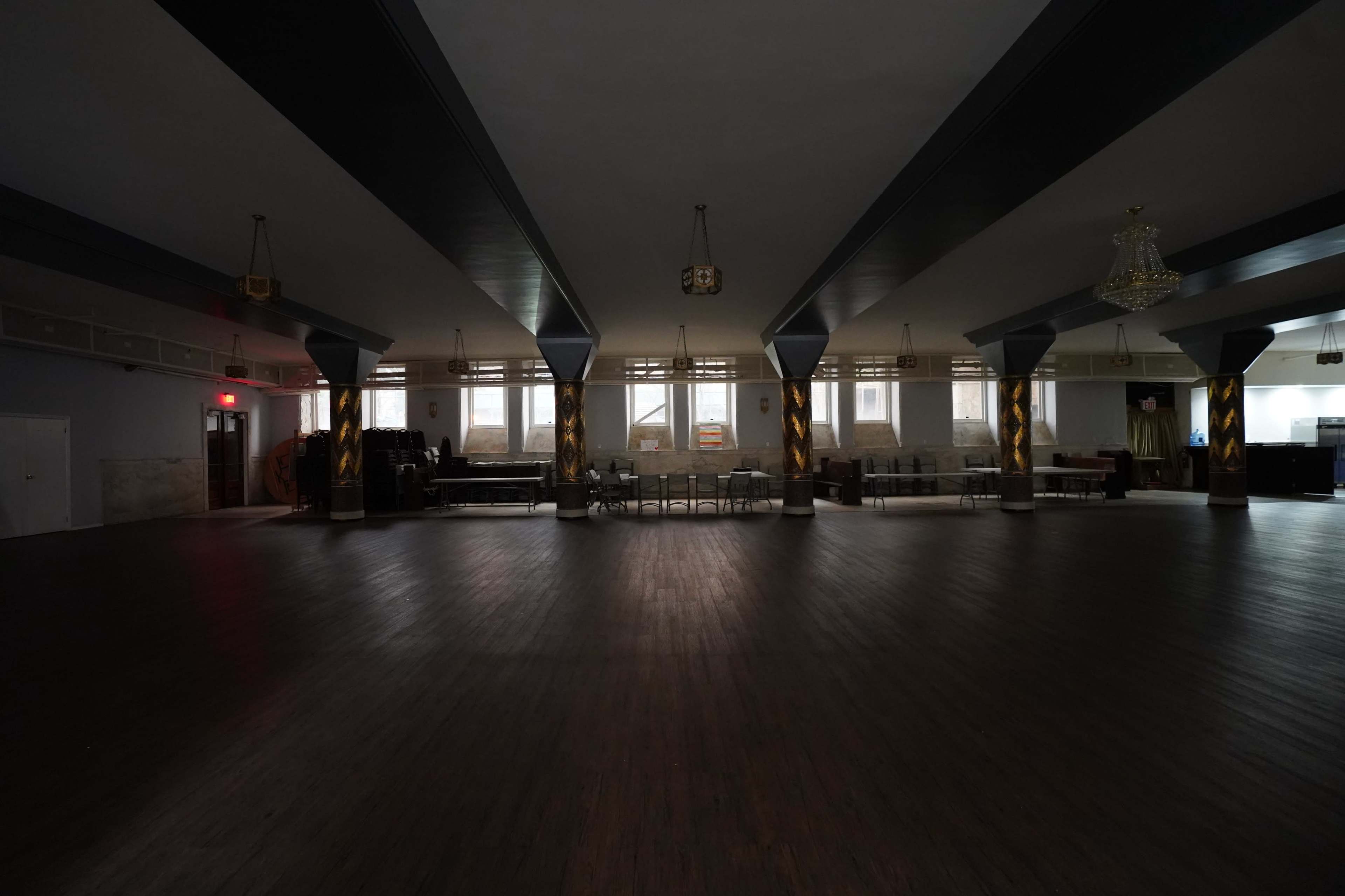 A spacious, dimly lit room with a wooden floor, high ceilings, and decorative columns, featuring various furniture items placed around the edges.