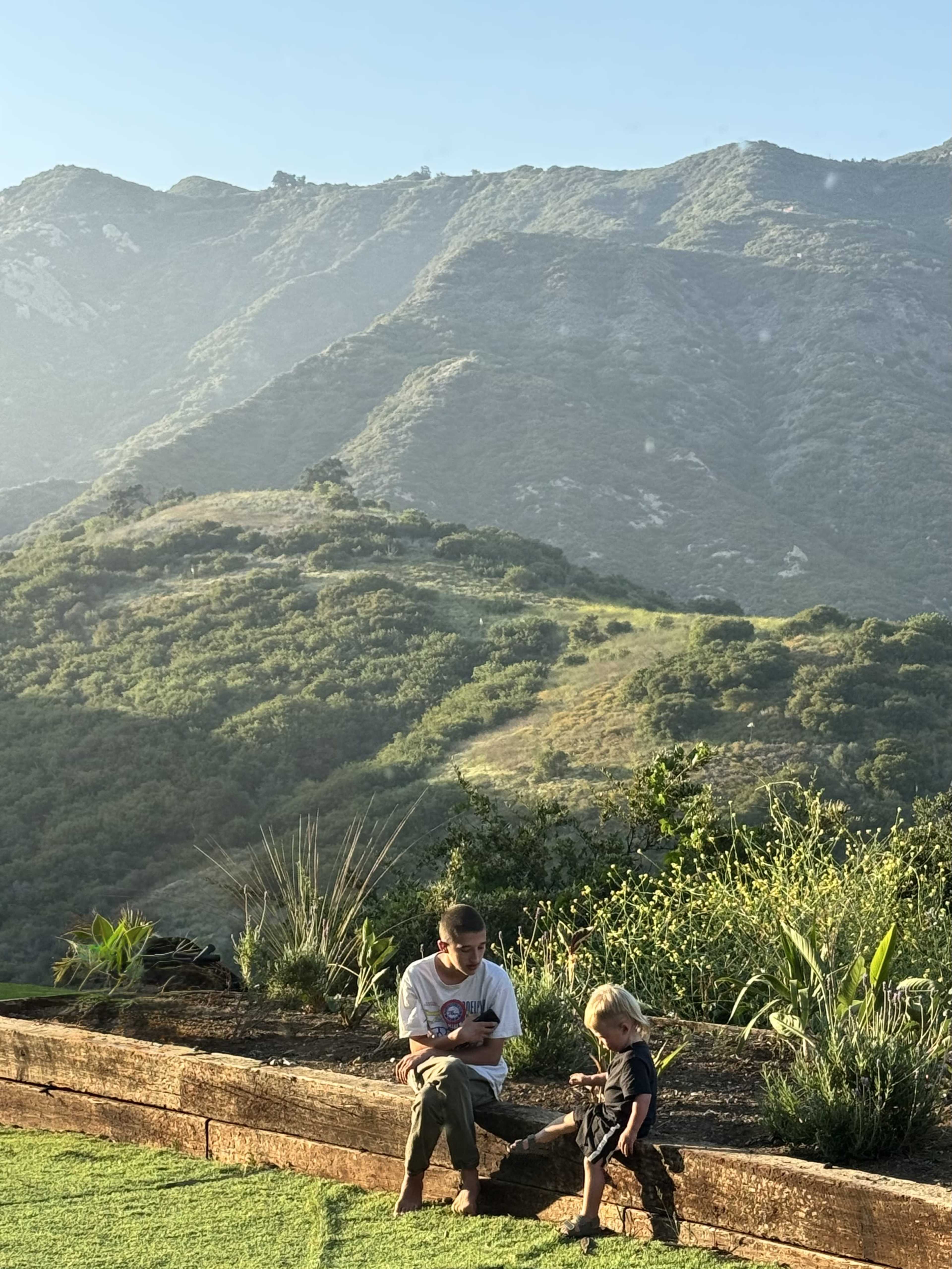 A child and an adult sit on a wooden bench in front of a grassy hillside with mountains in the background.