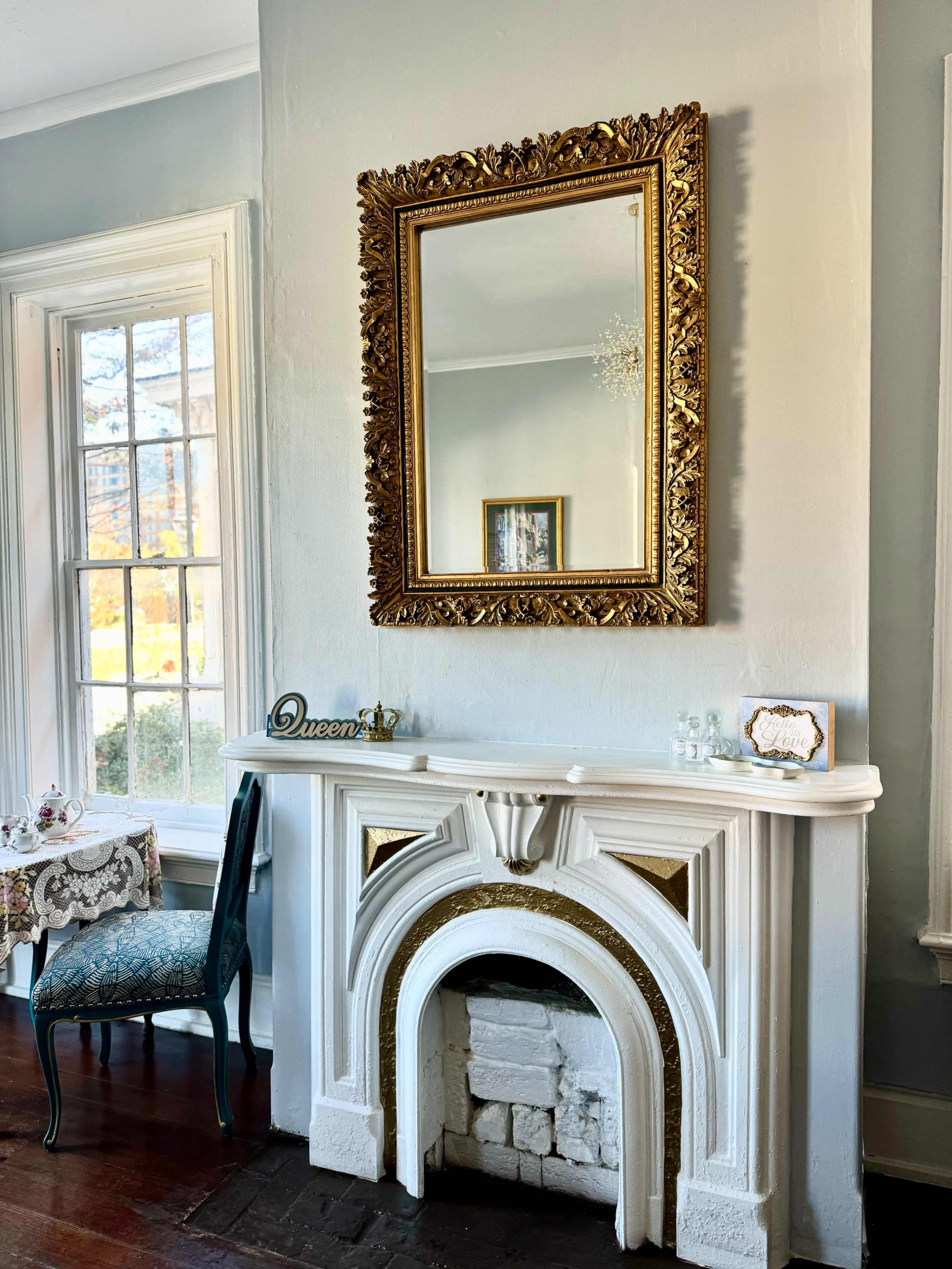 The image shows a decorative fireplace with a gold-framed mirror above it, alongside a blue chair and a small table adorned with a lace tablecloth.