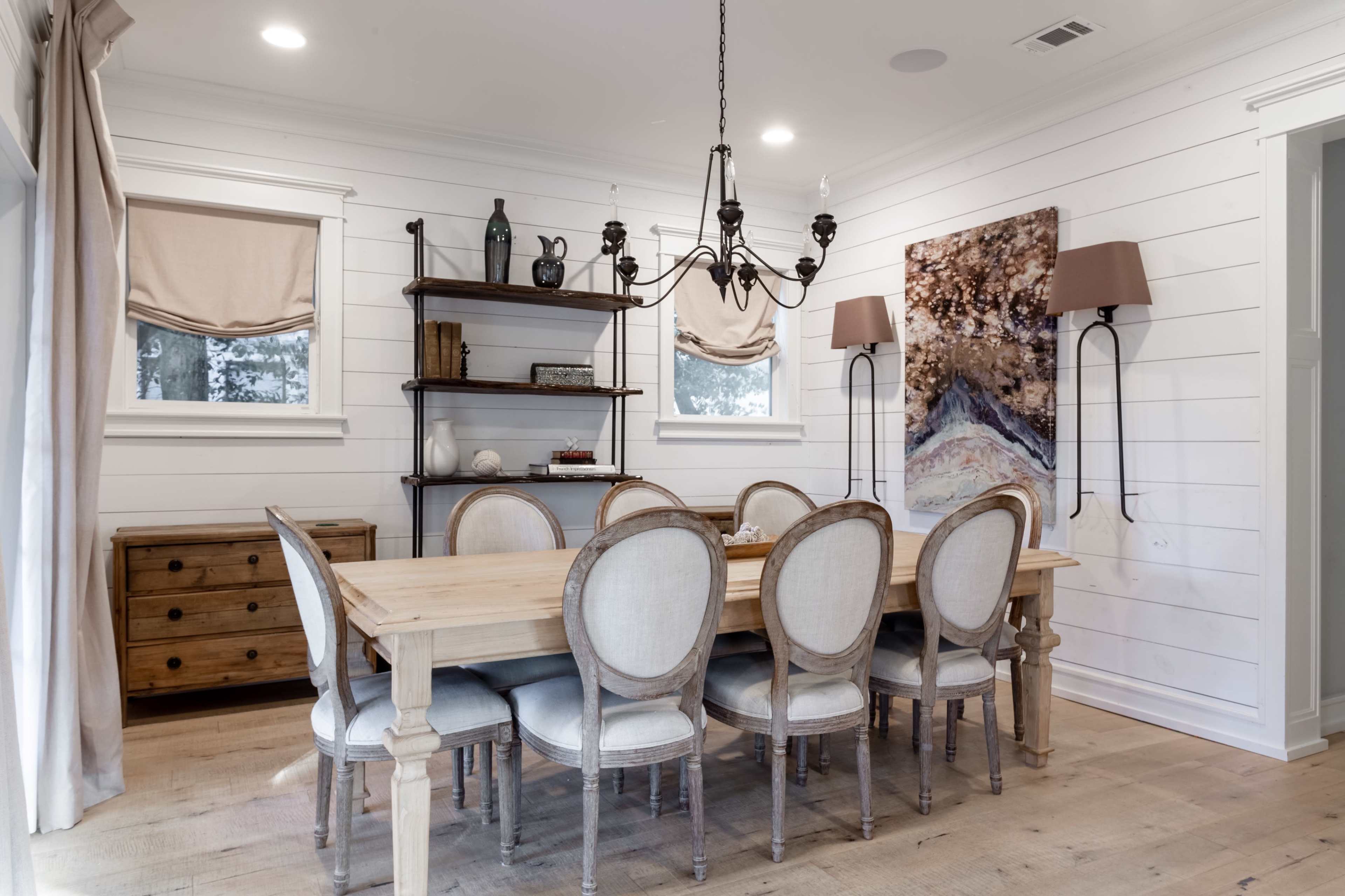 A dining room with a wooden table surrounded by eight upholstered chairs, a bookshelf, and wall-mounted lighting.
