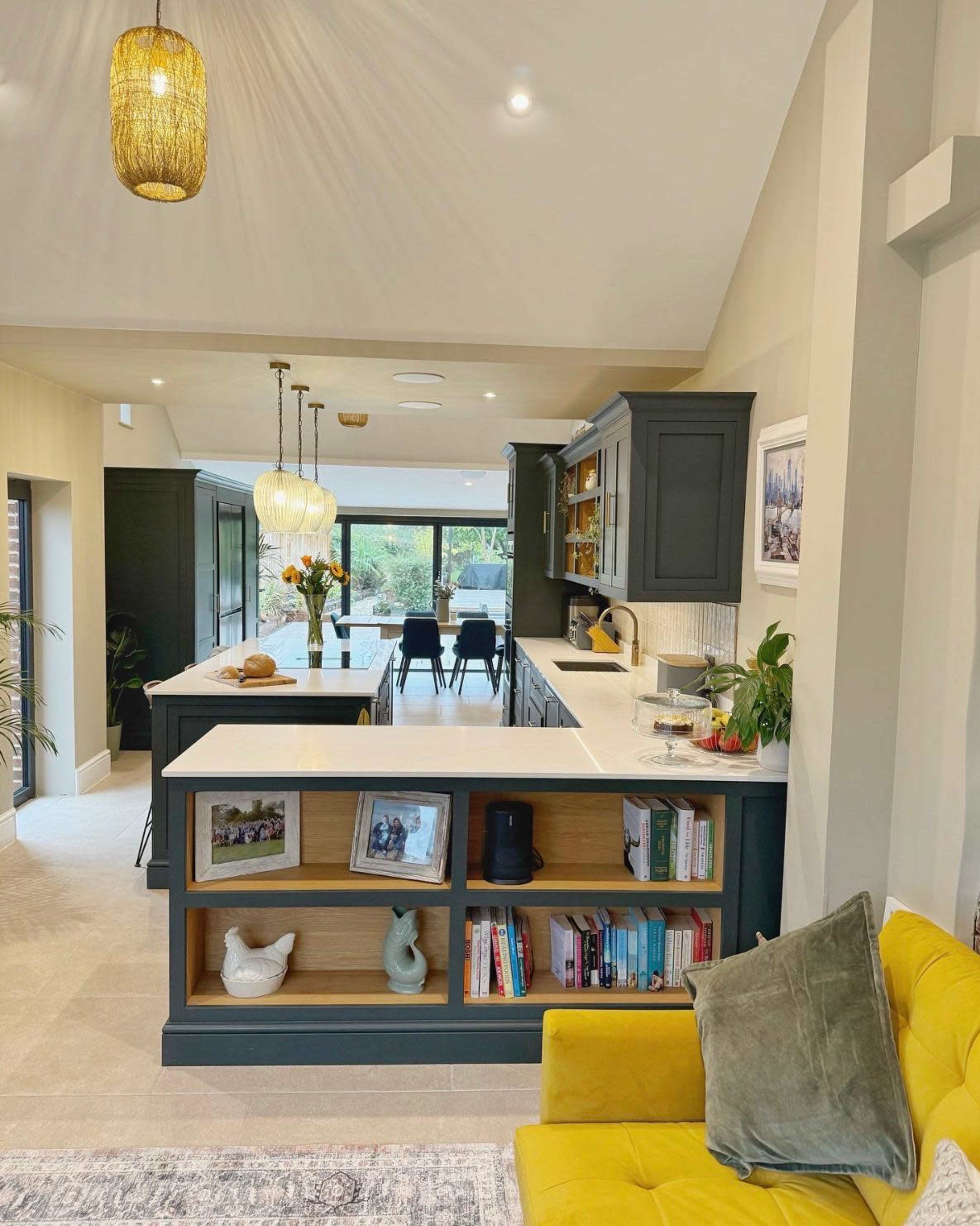 The image shows a modern open-concept kitchen and living area featuring a breakfast bar, shelves with books and decorations, and a dining table in the background.