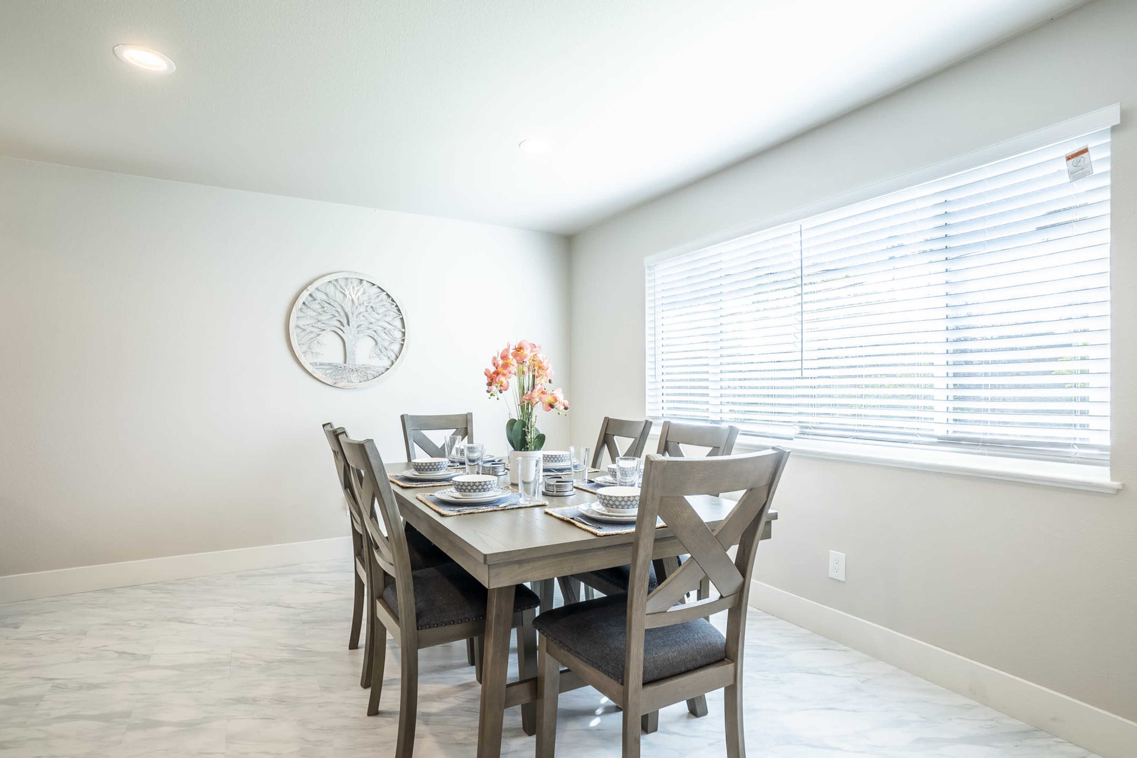 A dining area features a wooden table set for six, with floral centerpieces and a window covered in blinds.