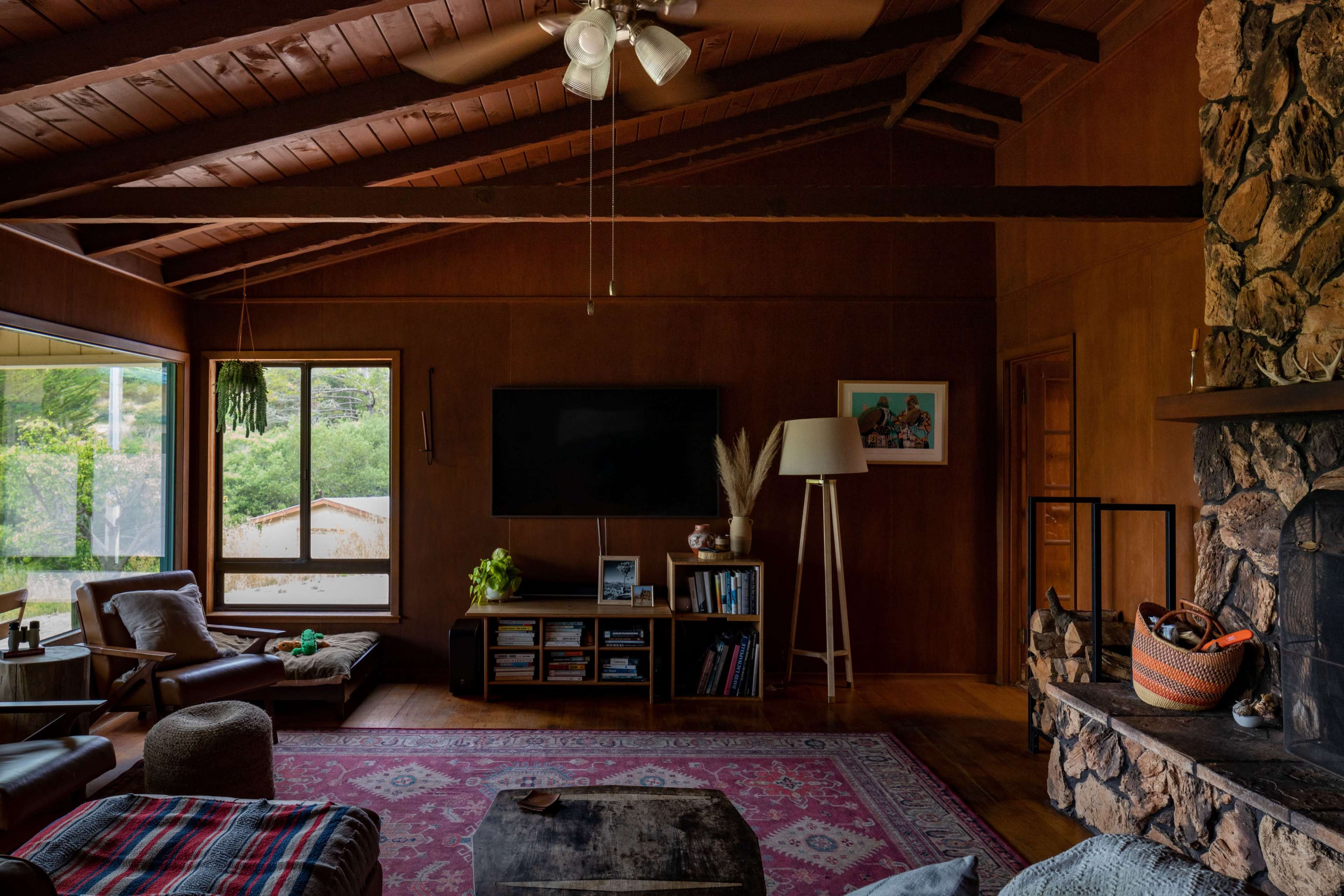 A living room with a stone fireplace, a television mounted on the wall, and large windows allowing natural light to enter.