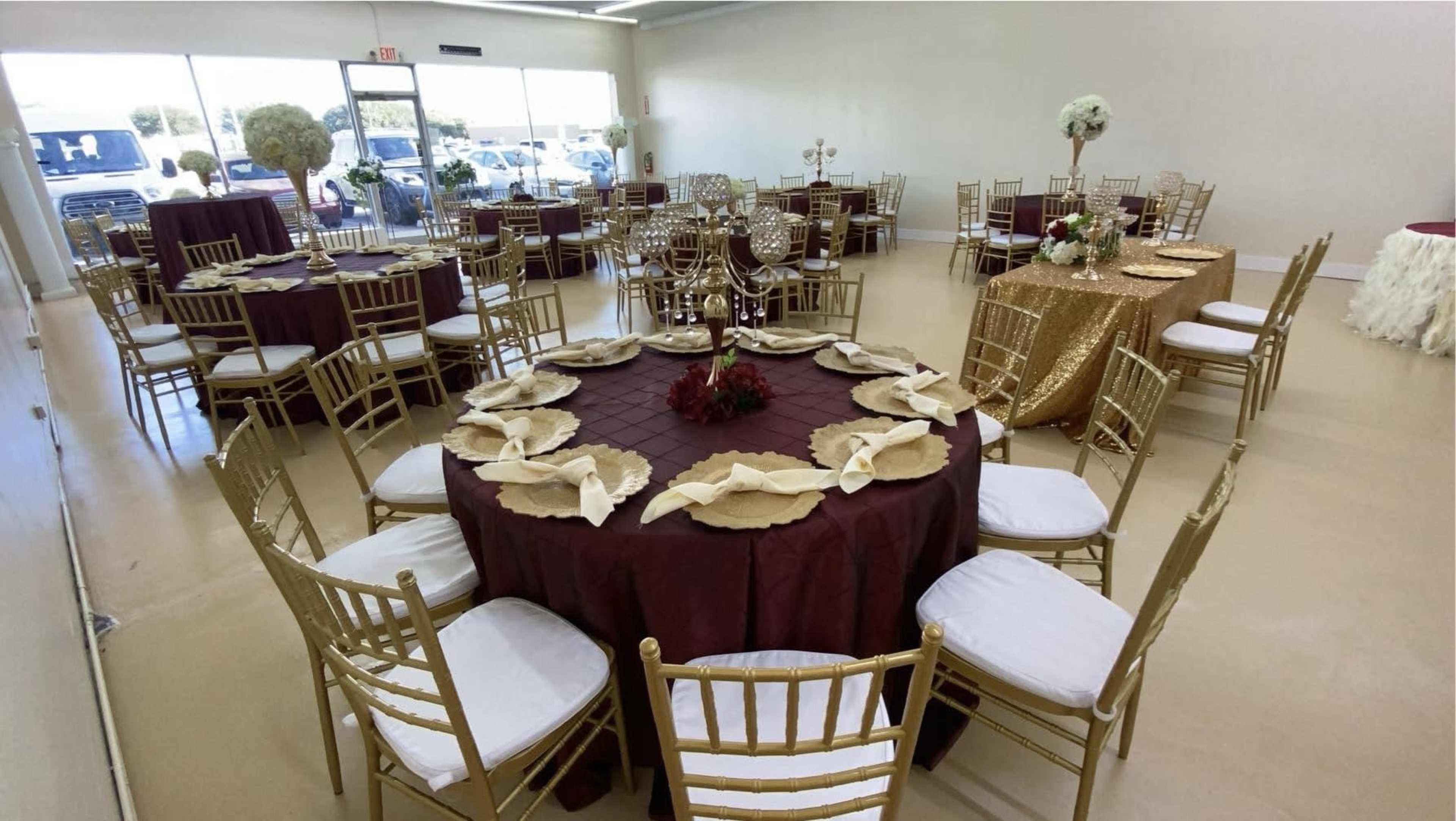 A spacious event room is set up with round tables covered in burgundy and gold linens, surrounded by gold chairs, and decorated with floral centerpieces.