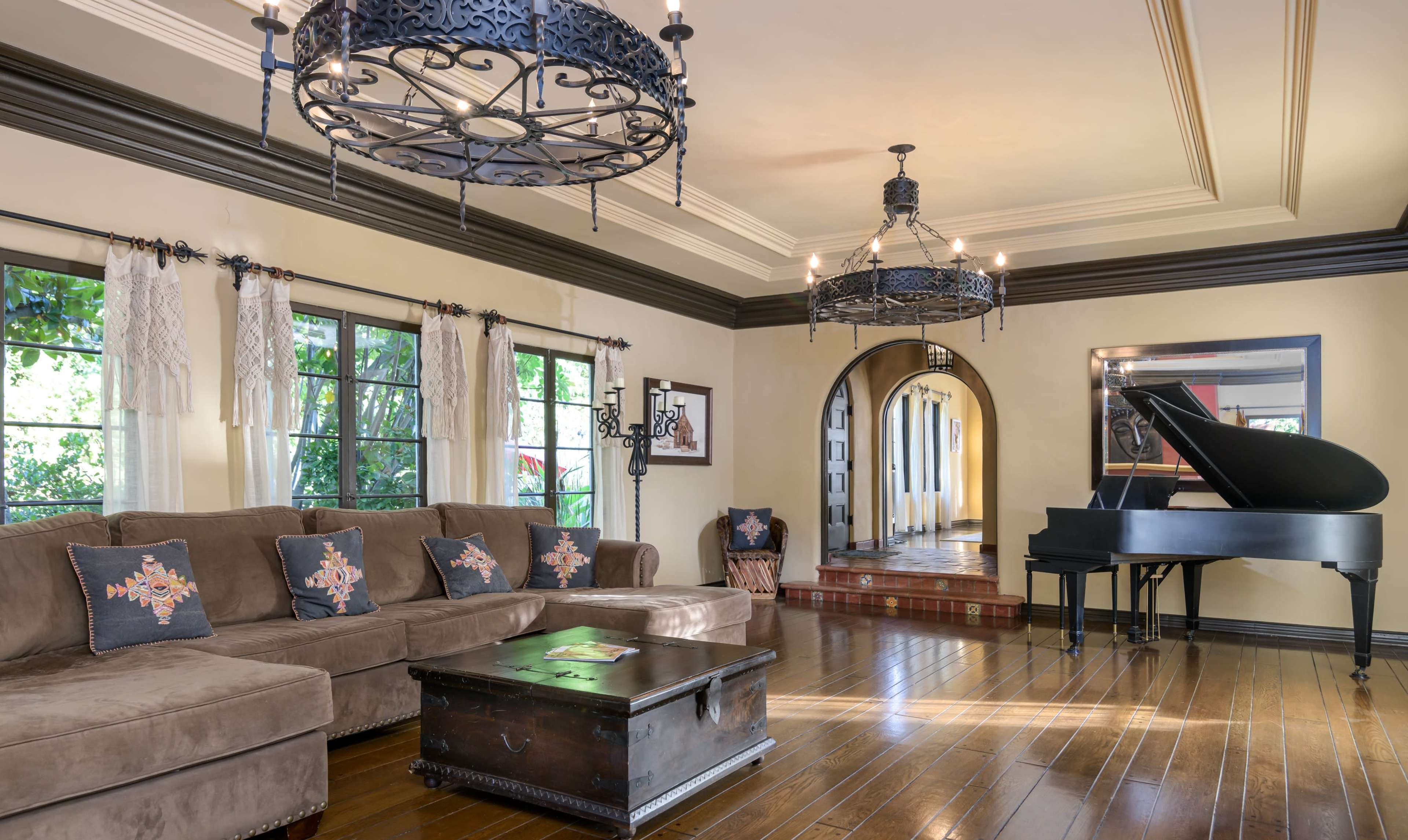 A spacious living room features a brown sectional sofa, a black grand piano, and two ornate chandeliers hanging from the ceiling.