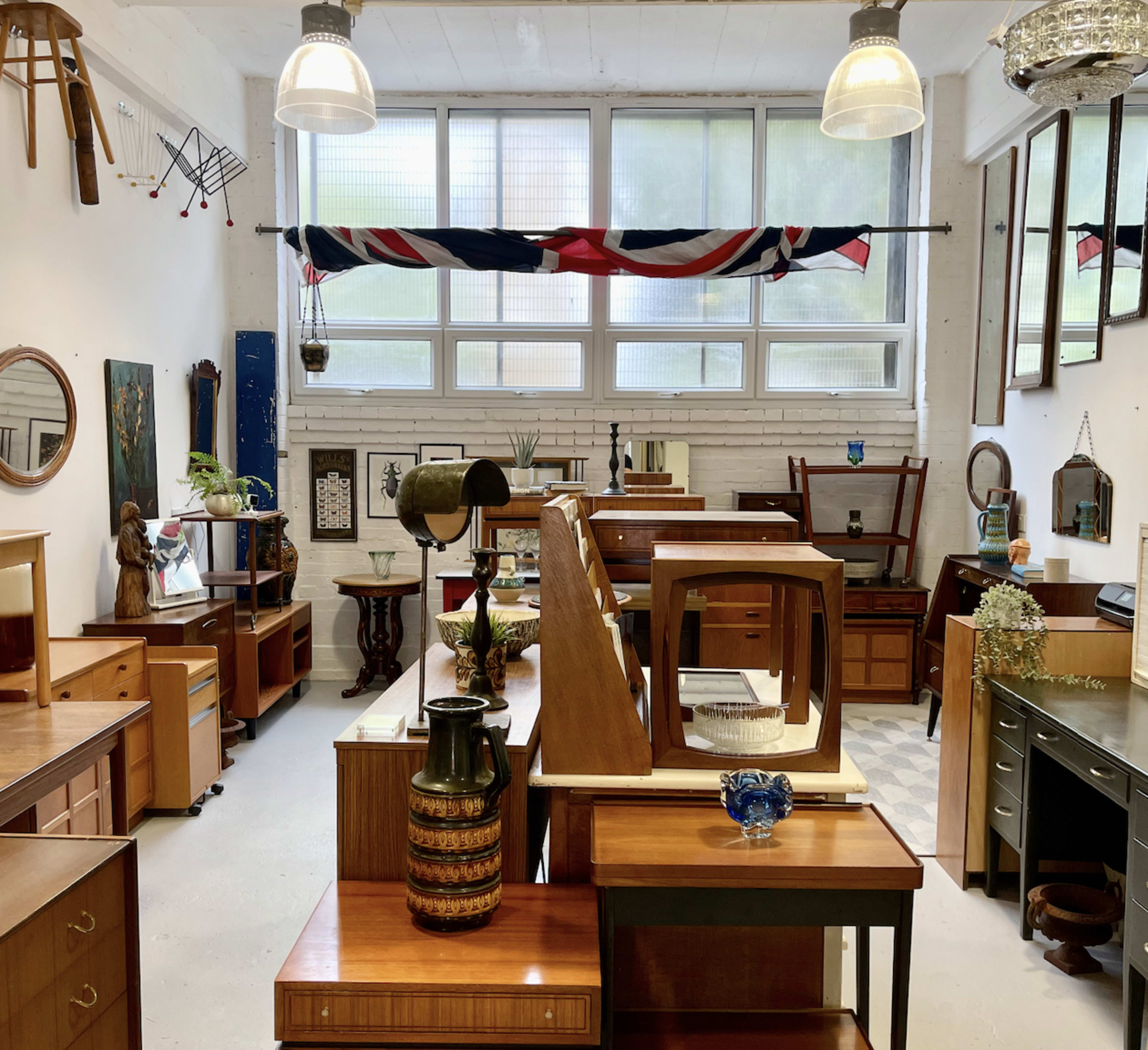 A spacious room filled with various wooden furniture pieces, including tables, cabinets, and decorative items, under large windows with a British flag hanging across the top.