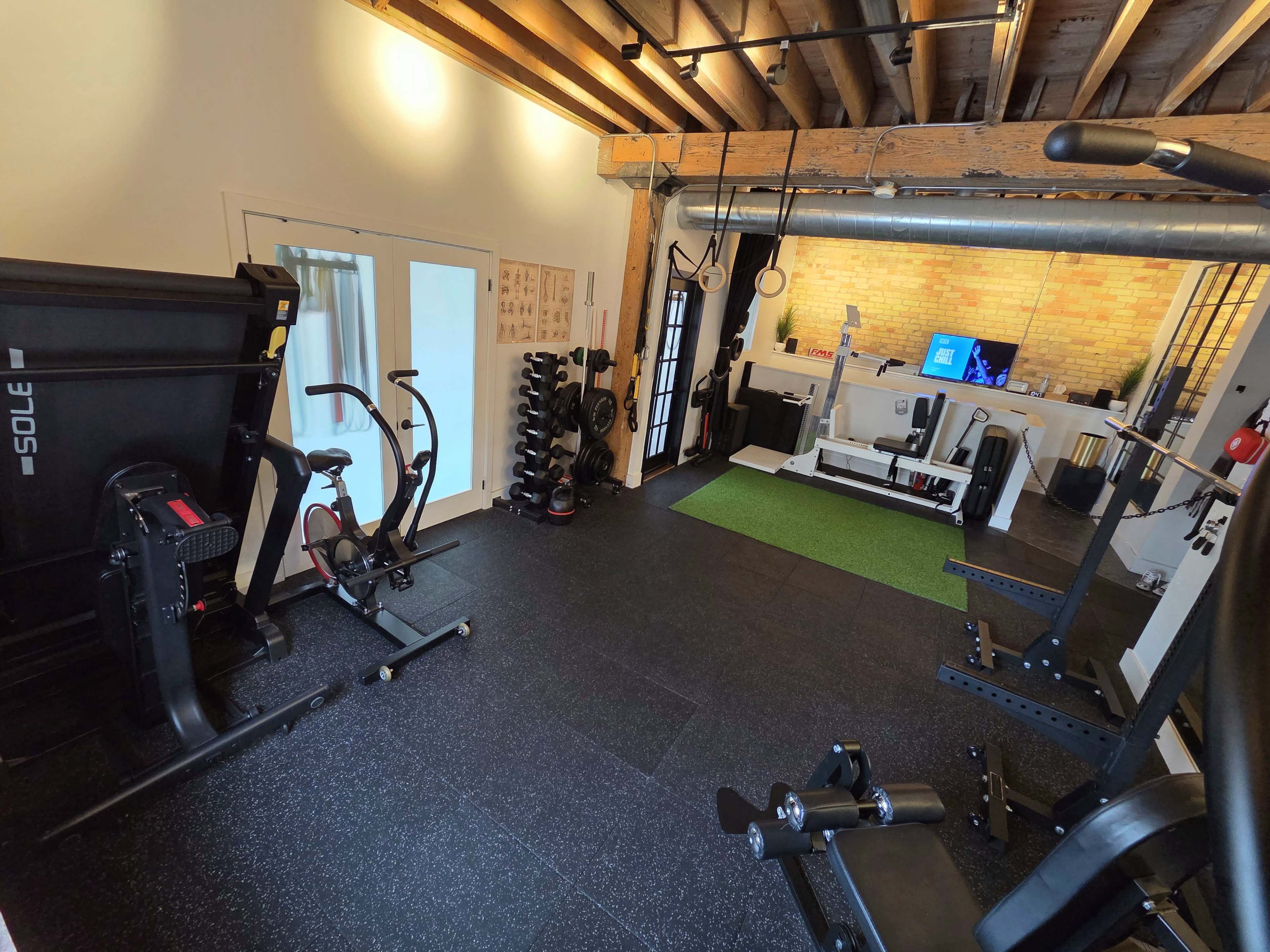 The image shows a well-equipped home gym featuring cardio and weight training equipment on black rubber flooring.
