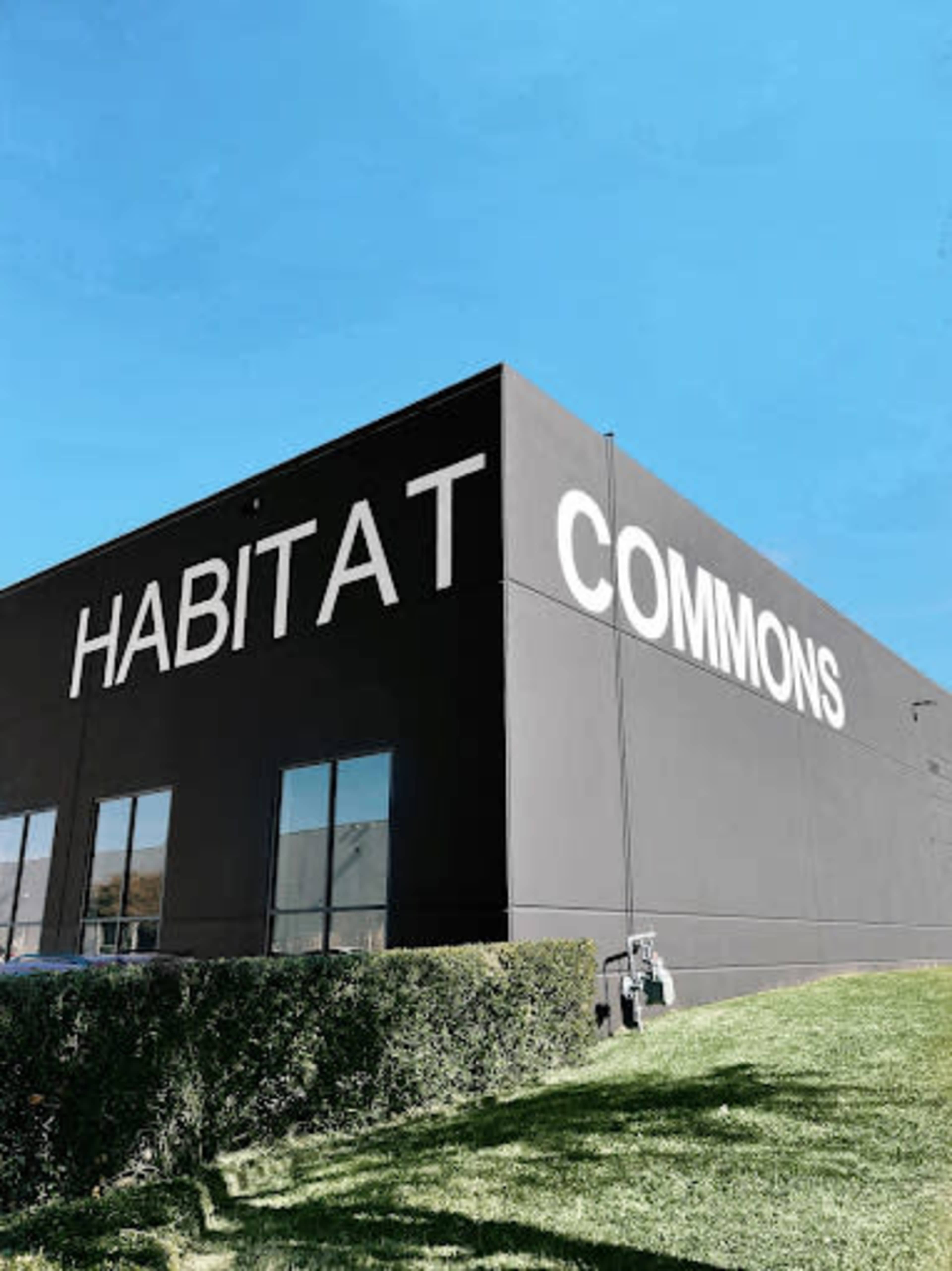 The image shows a large black building with the words "HABITAT COMMONS" prominently displayed on its side under a clear blue sky.