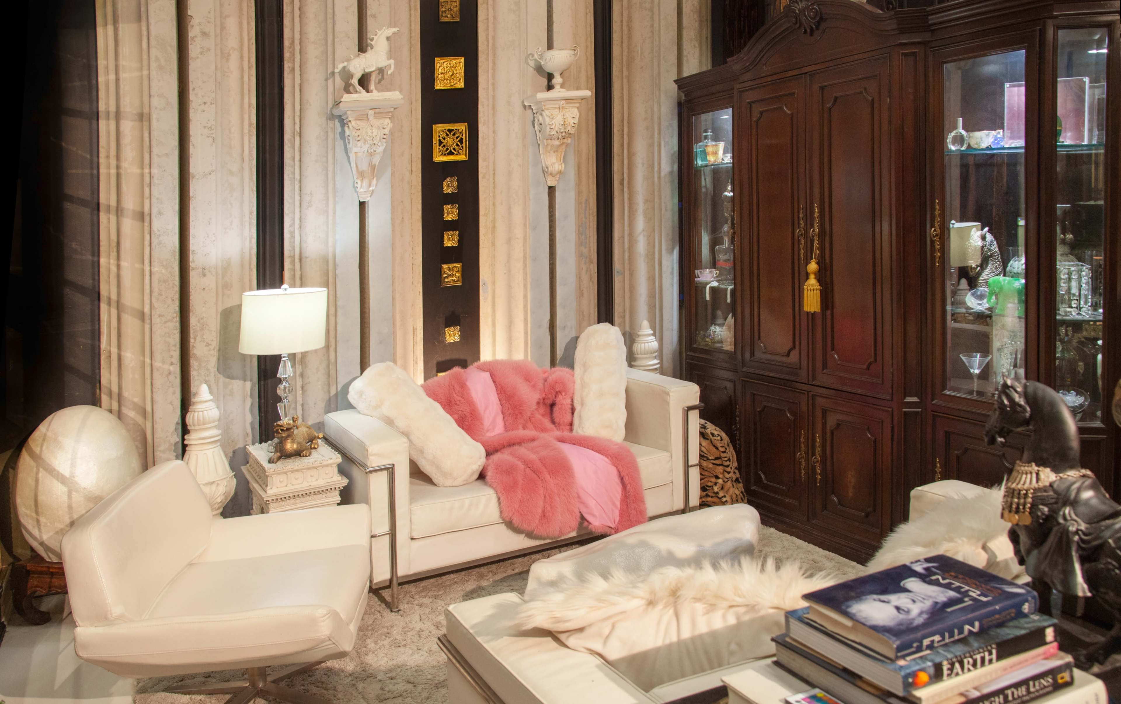 The image features a stylish living room with a white leather sofa, a pink throw blanket, a glass cabinet displaying decorative items, and a coffee table topped with books.