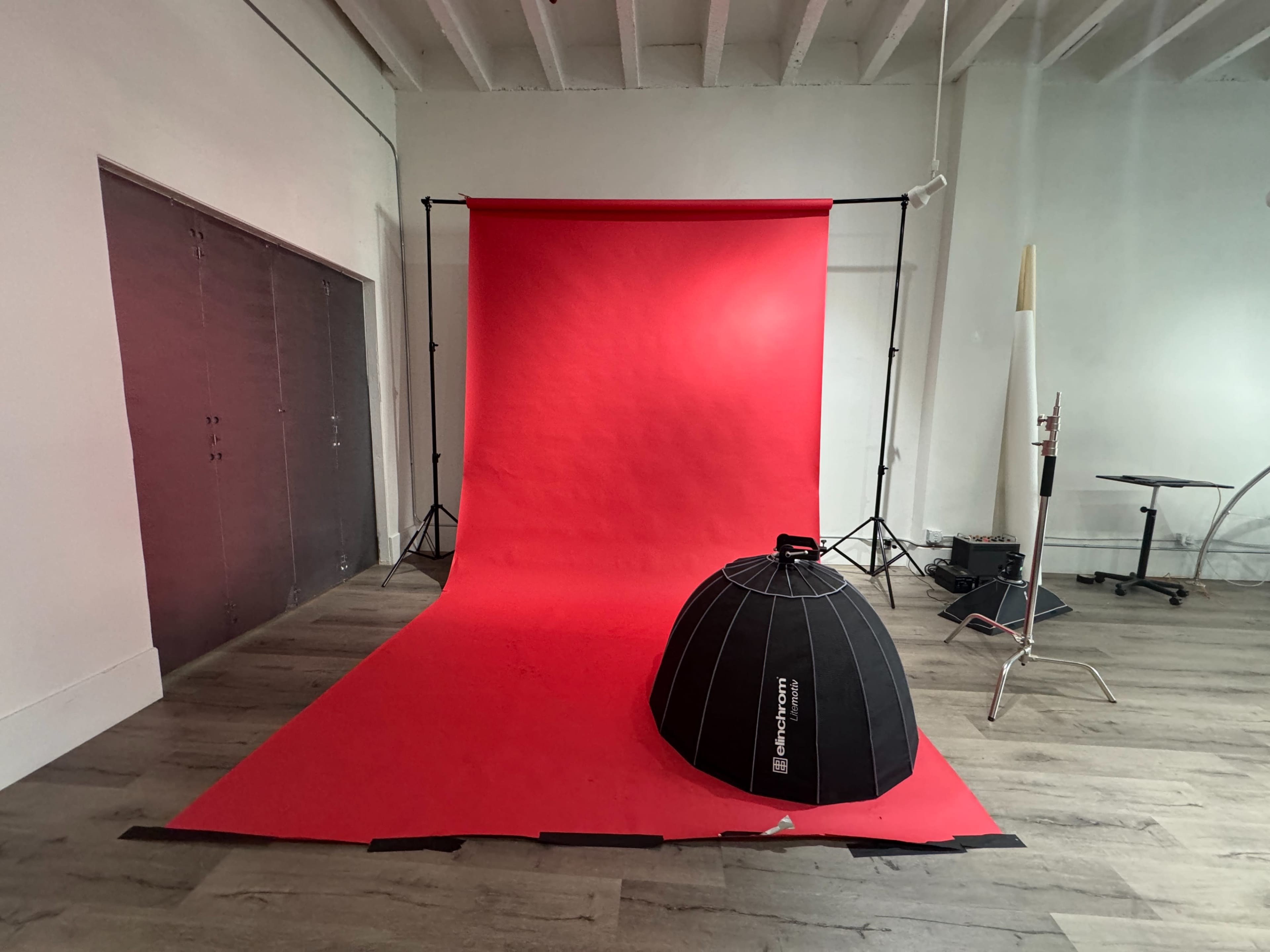 The image shows a photography studio with a red backdrop, a black light modifier on the floor, and various studio equipment visible.