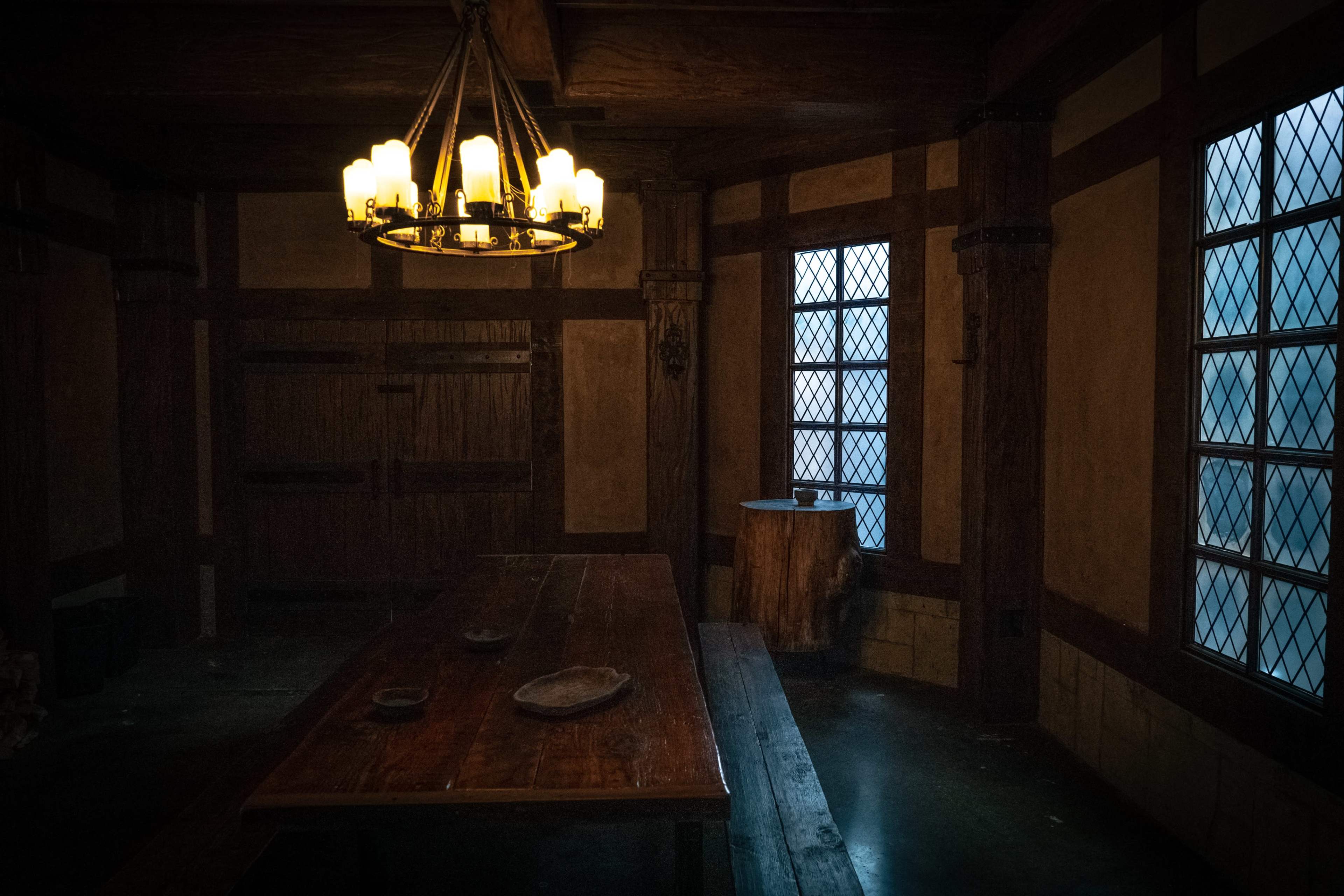 The image depicts a dimly lit, rustic room with wooden beams, a long table, and two windows featuring diamond-patterned glass.
