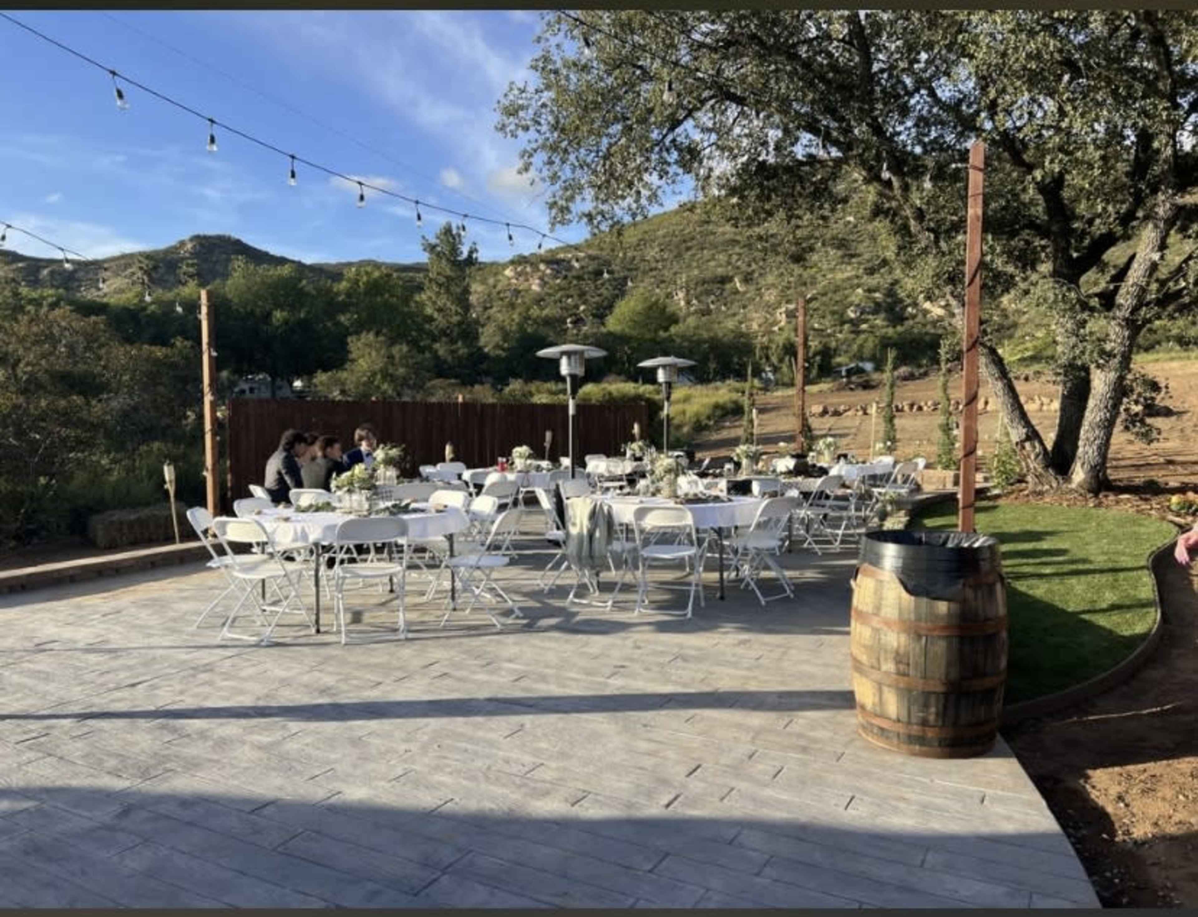 The scene features a patio area set with several tables and chairs under string lights, surrounded by trees and hills.