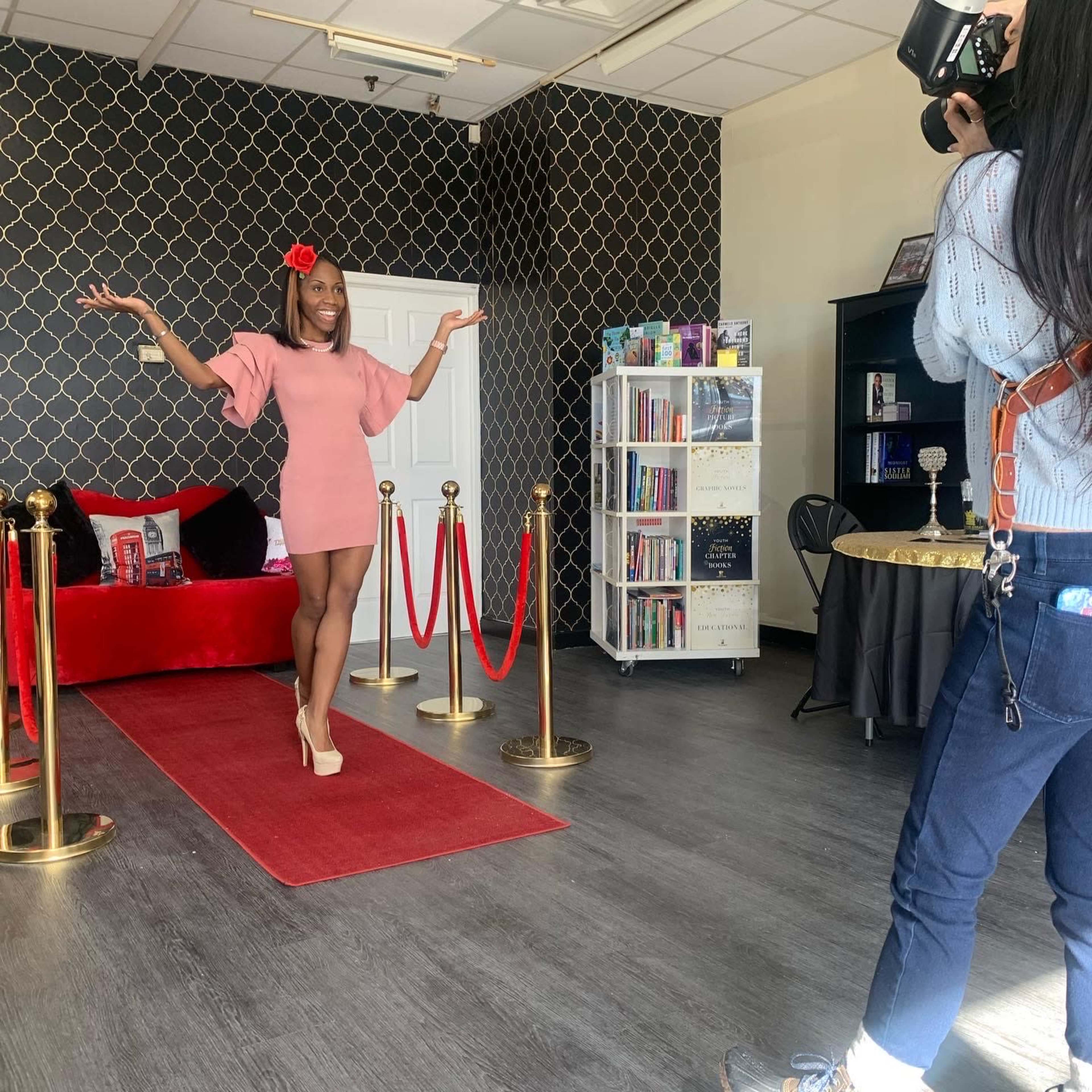 A woman in a pink dress poses with her arms raised in a studio setting while a photographer captures the moment.
