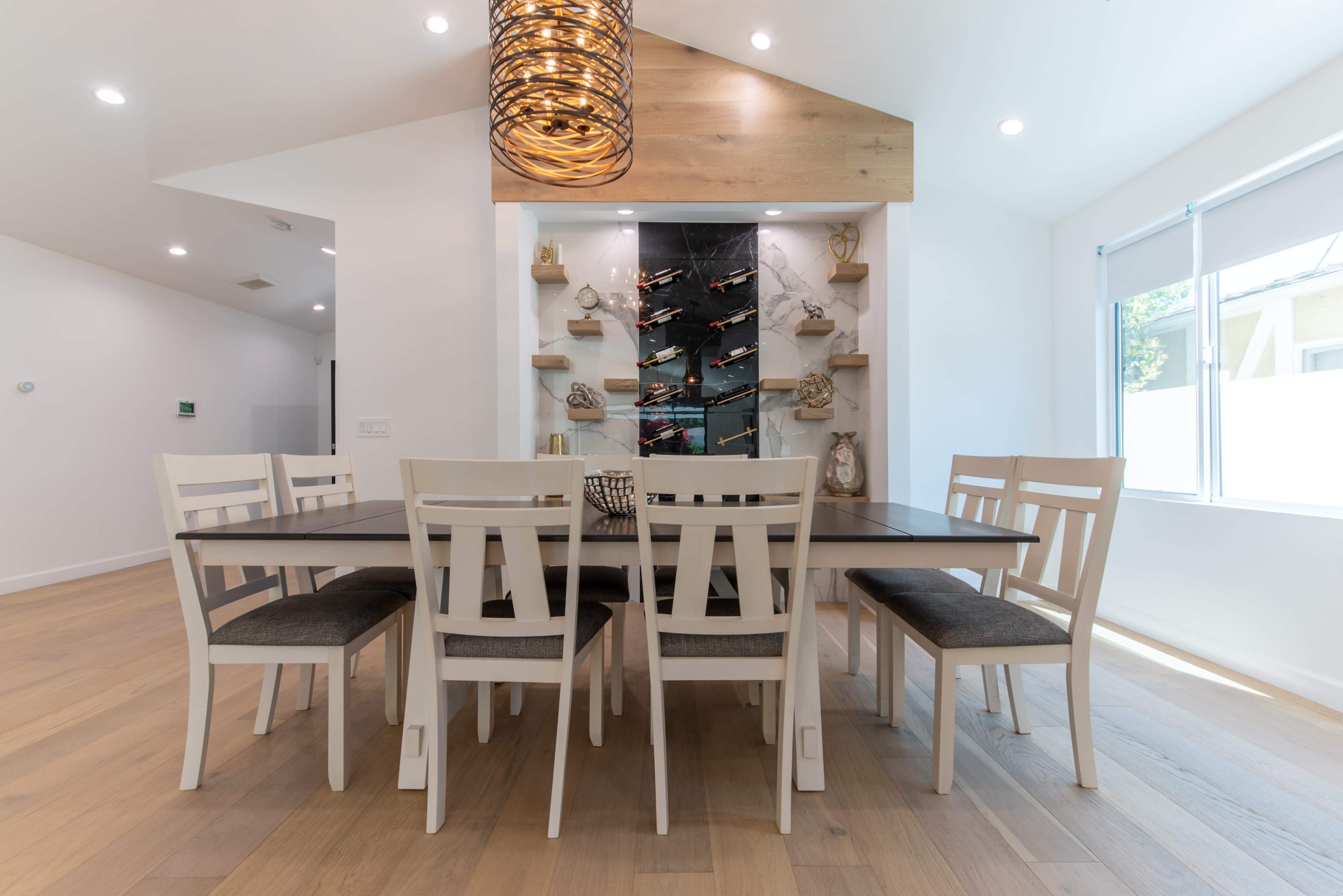 A modern dining area features a large table surrounded by six chairs, with a decorative wall display in the background and ample natural light coming through the windows.