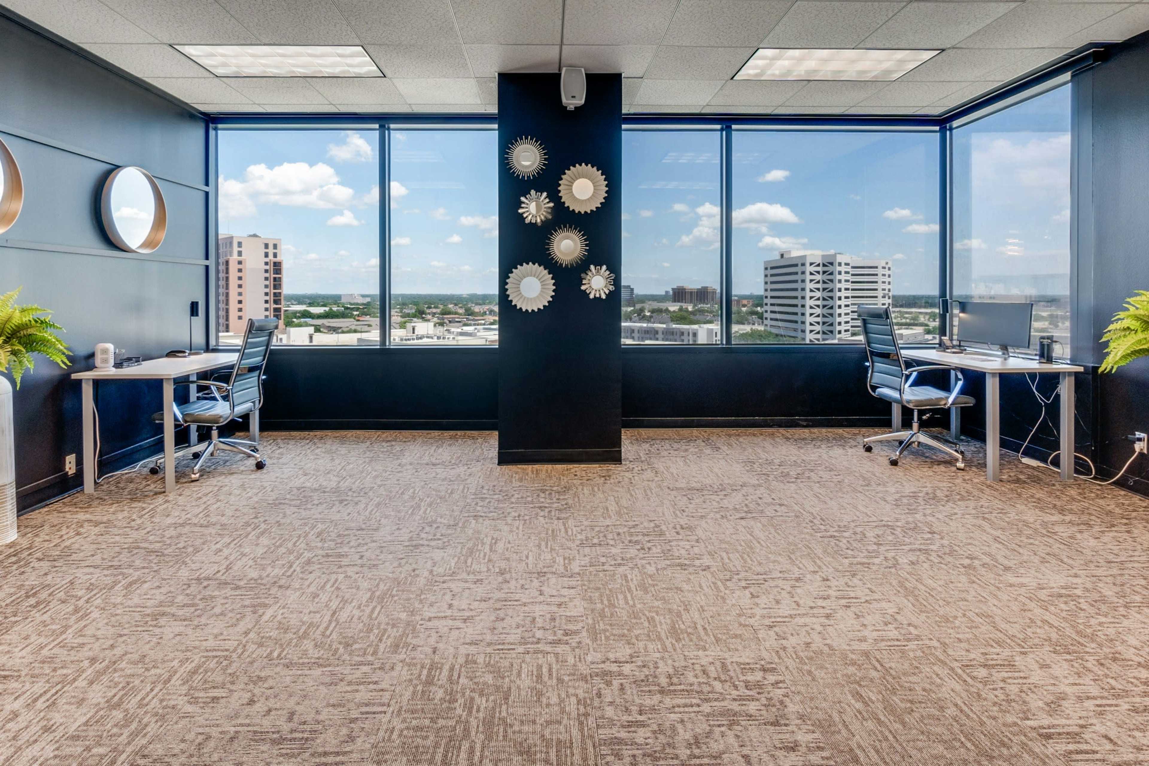 The image shows a modern office space with two desks facing large windows that offer a view of a city landscape.