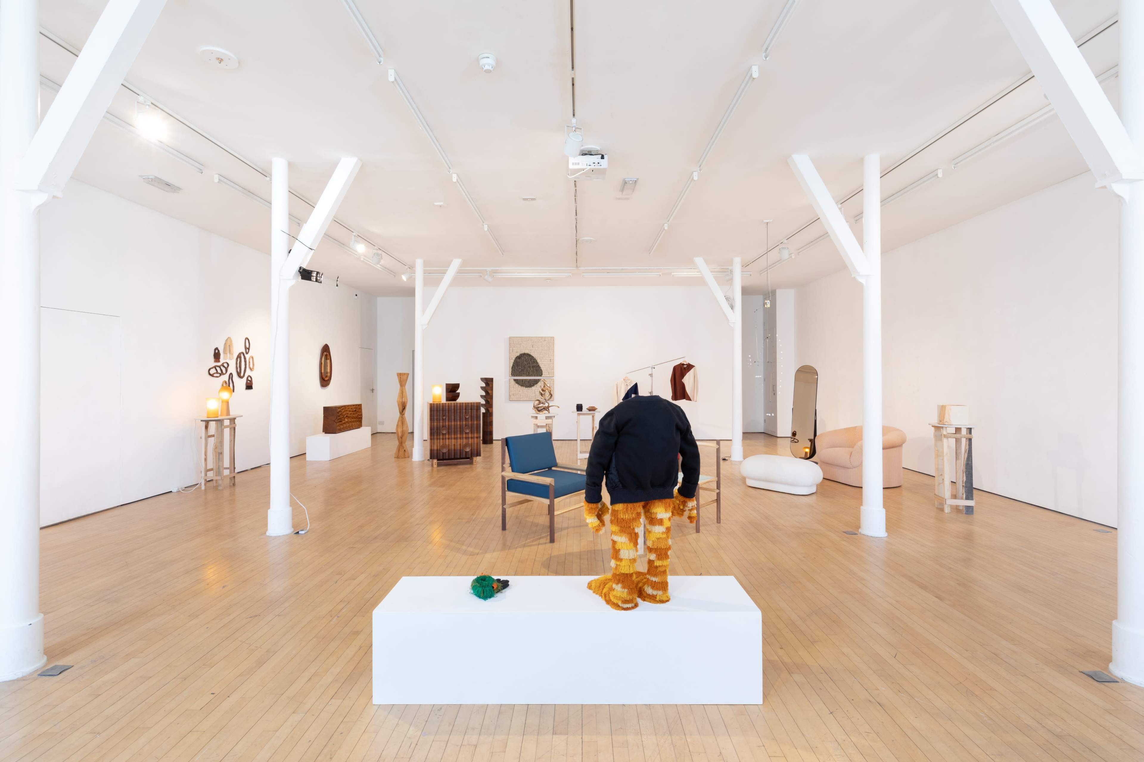 A contemporary art gallery featuring various sculptures and furniture pieces arranged in a spacious, well-lit room with white walls and wooden flooring.