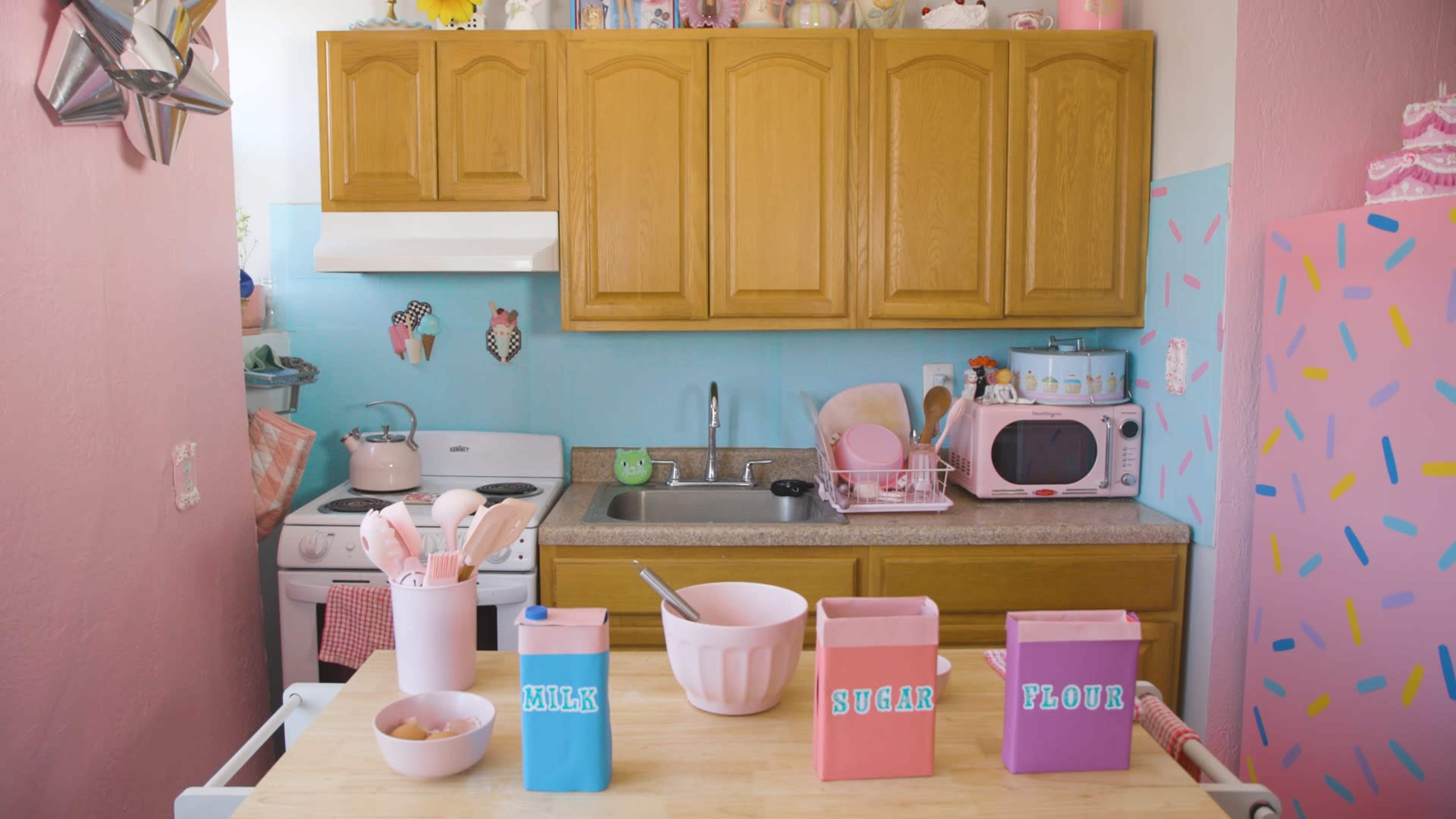 A kitchen with wooden cabinets and pastel-colored walls features a countertop with labeled containers for milk, sugar, and flour, along with cooking utensils and a mixing bowl.