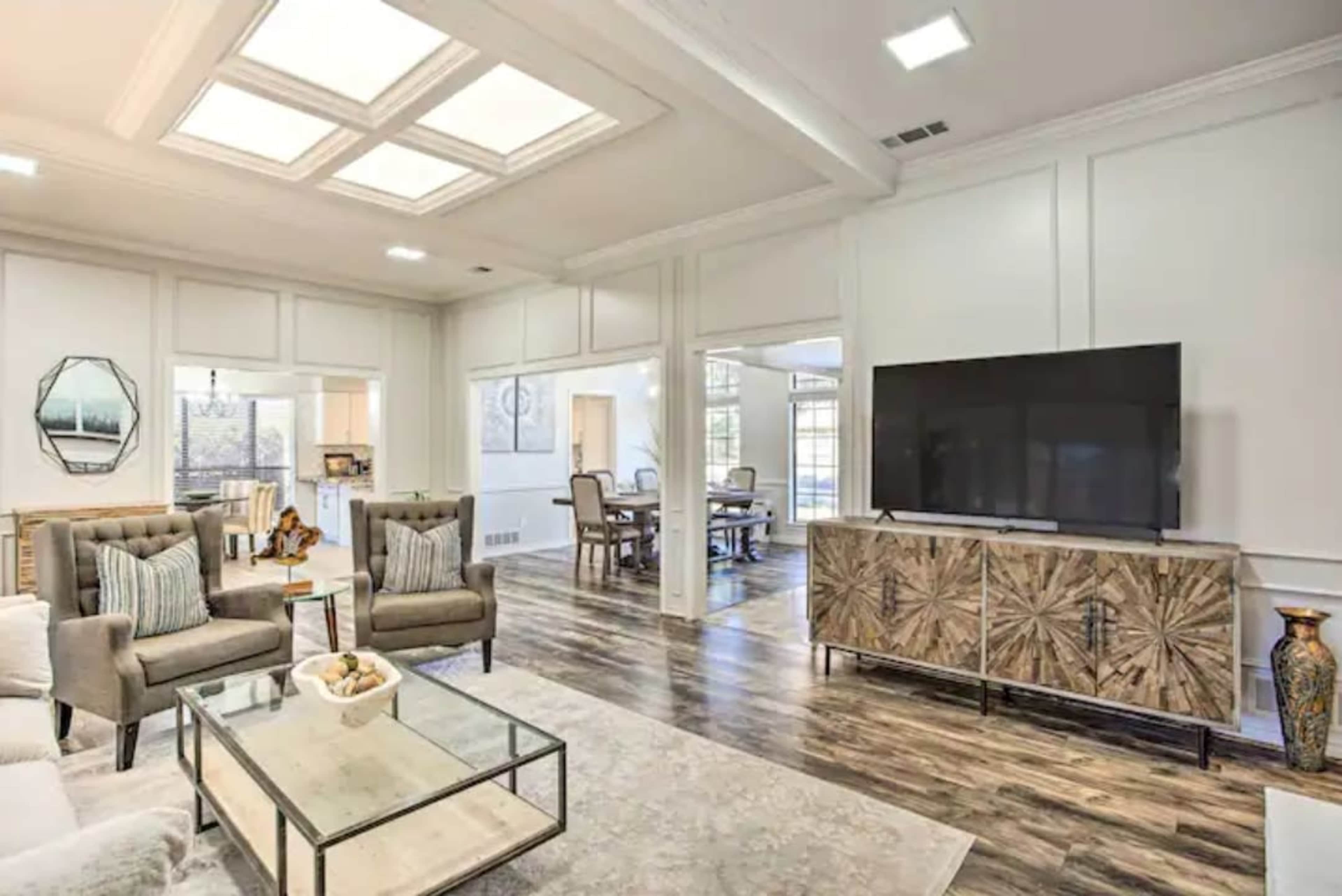 A modern living room with light-colored walls, a large television on a textured media console, two armchairs, and a glass coffee table, with an open view to another room.