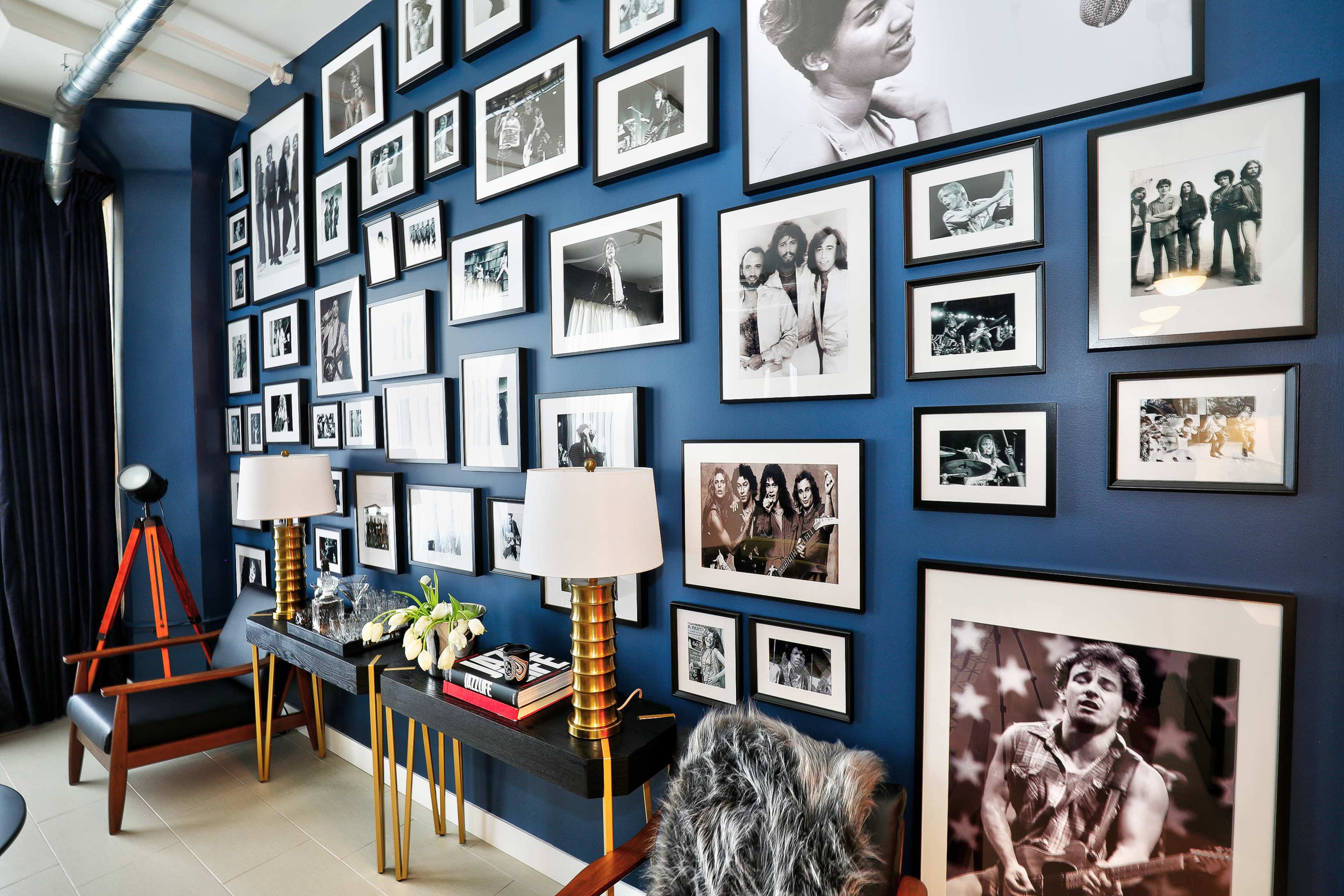 The image features a wall adorned with a diverse collection of black-and-white framed photographs, alongside two tables with lamps and decorative items set against a deep blue backdrop.