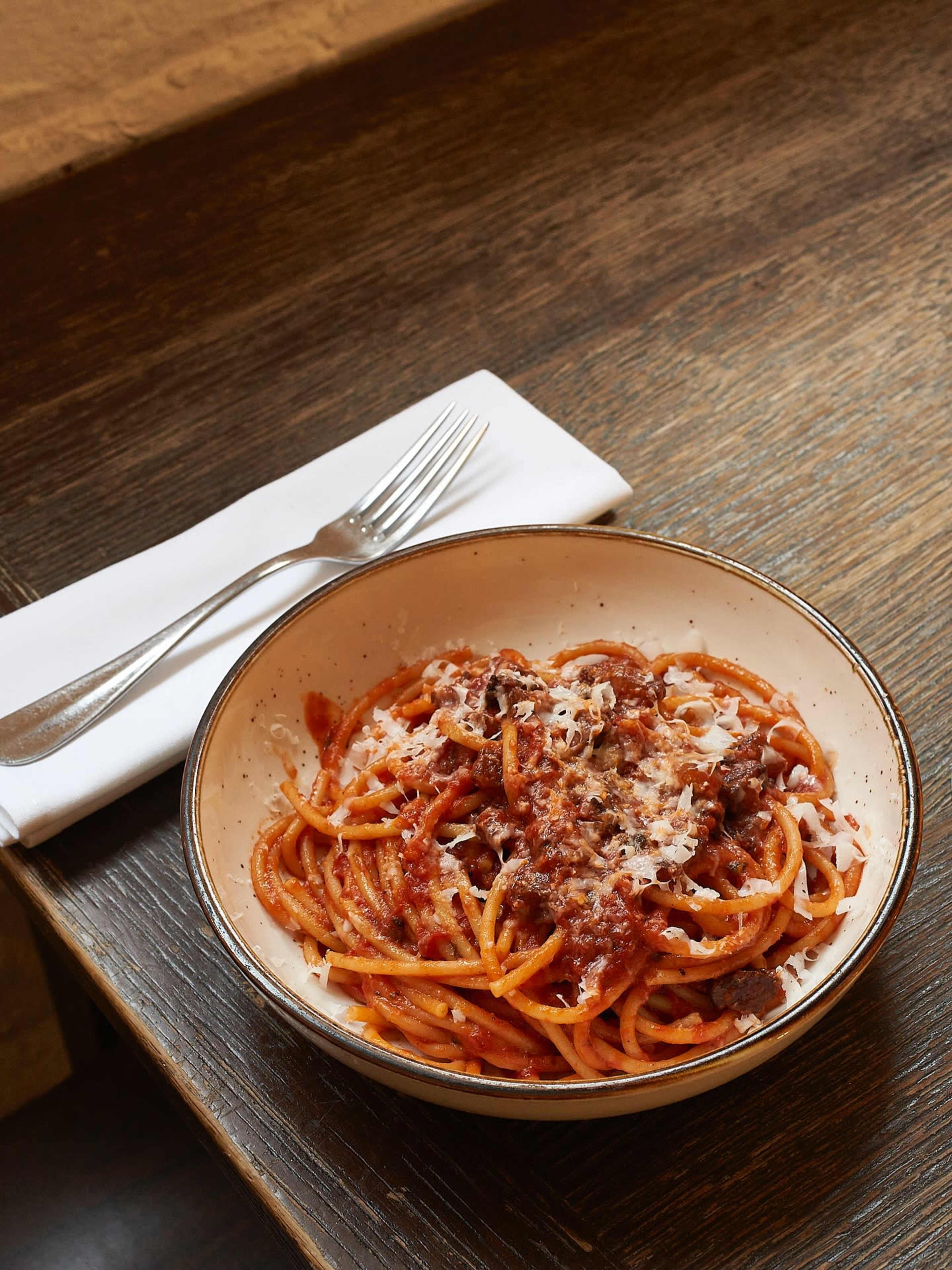 A bowl of spaghetti topped with meat sauce and grated cheese sits on a wooden table next to a folded napkin and a fork.