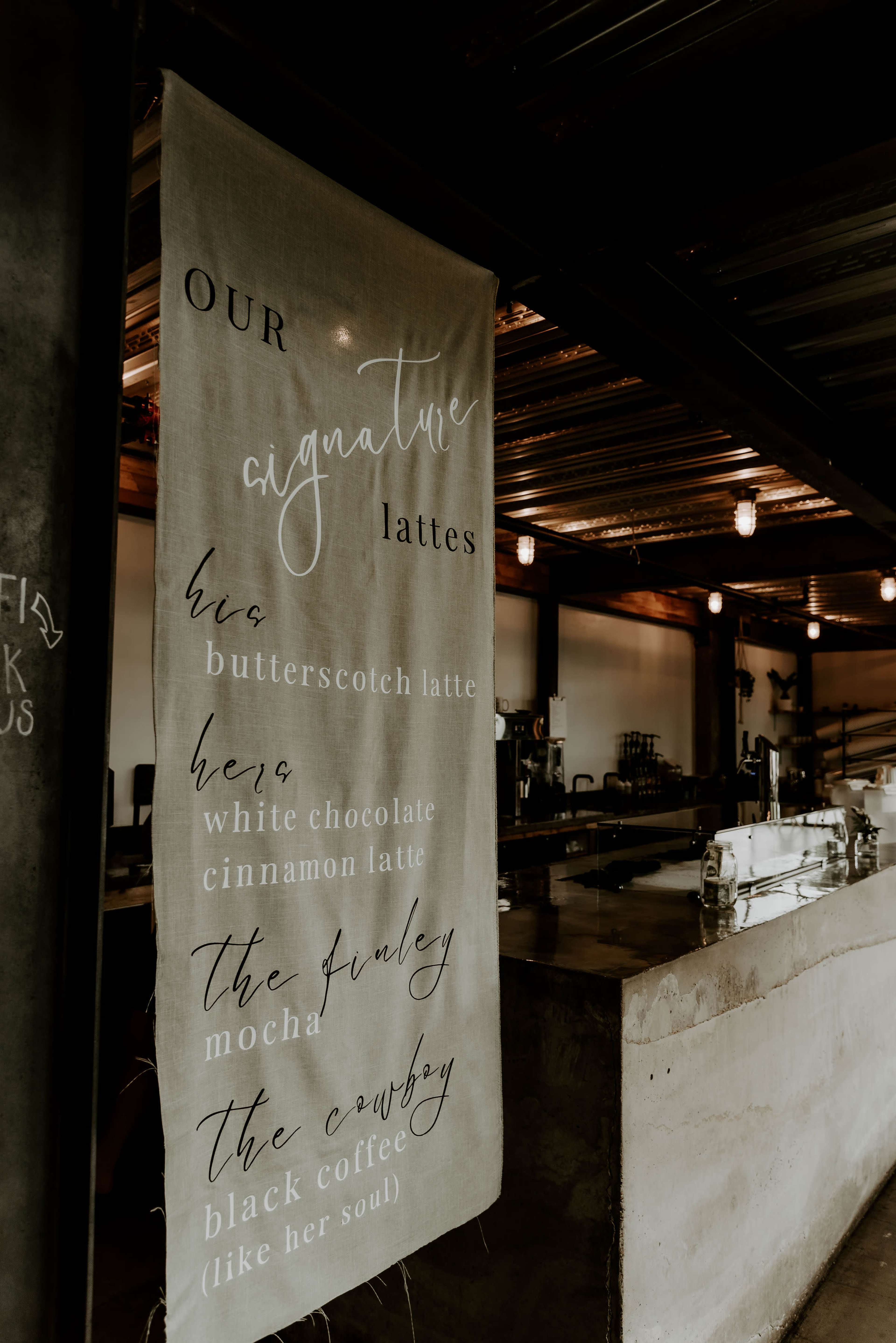 A fabric banner displays a list of signature lattes, including butterscotch, white chocolate cinnamon, and mocha, in a coffee shop setting.