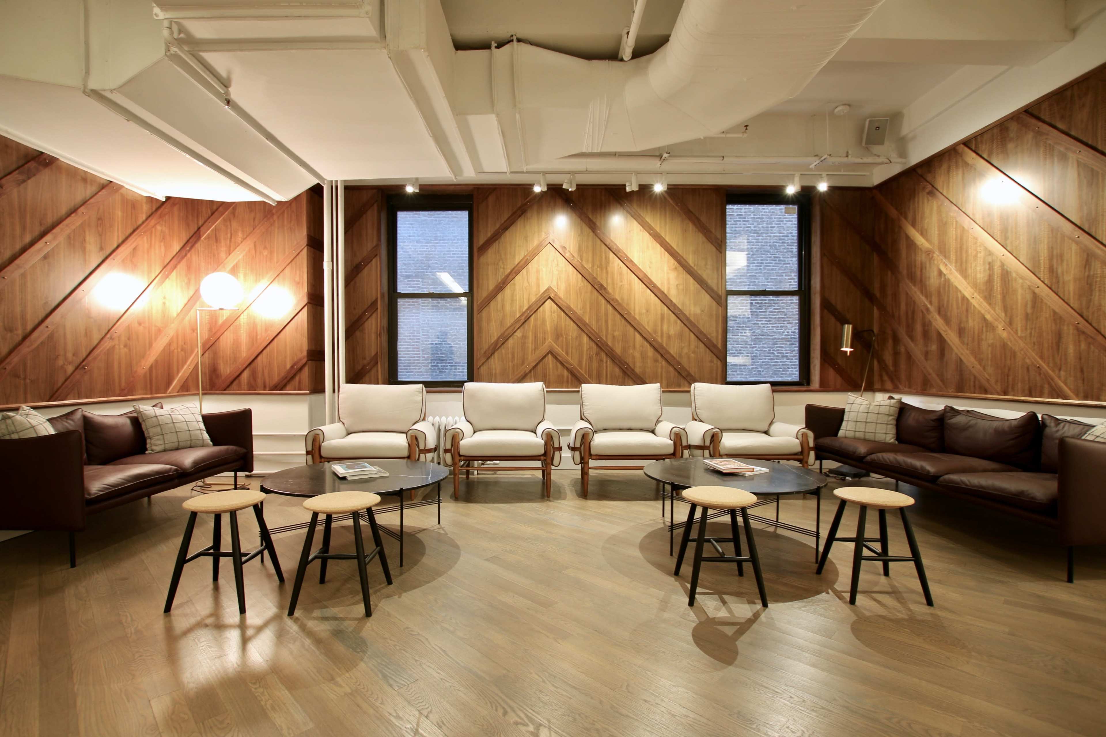 The image shows a stylish lounge area with wooden walls, a combination of white and dark brown seating, and round tables arranged in the center.