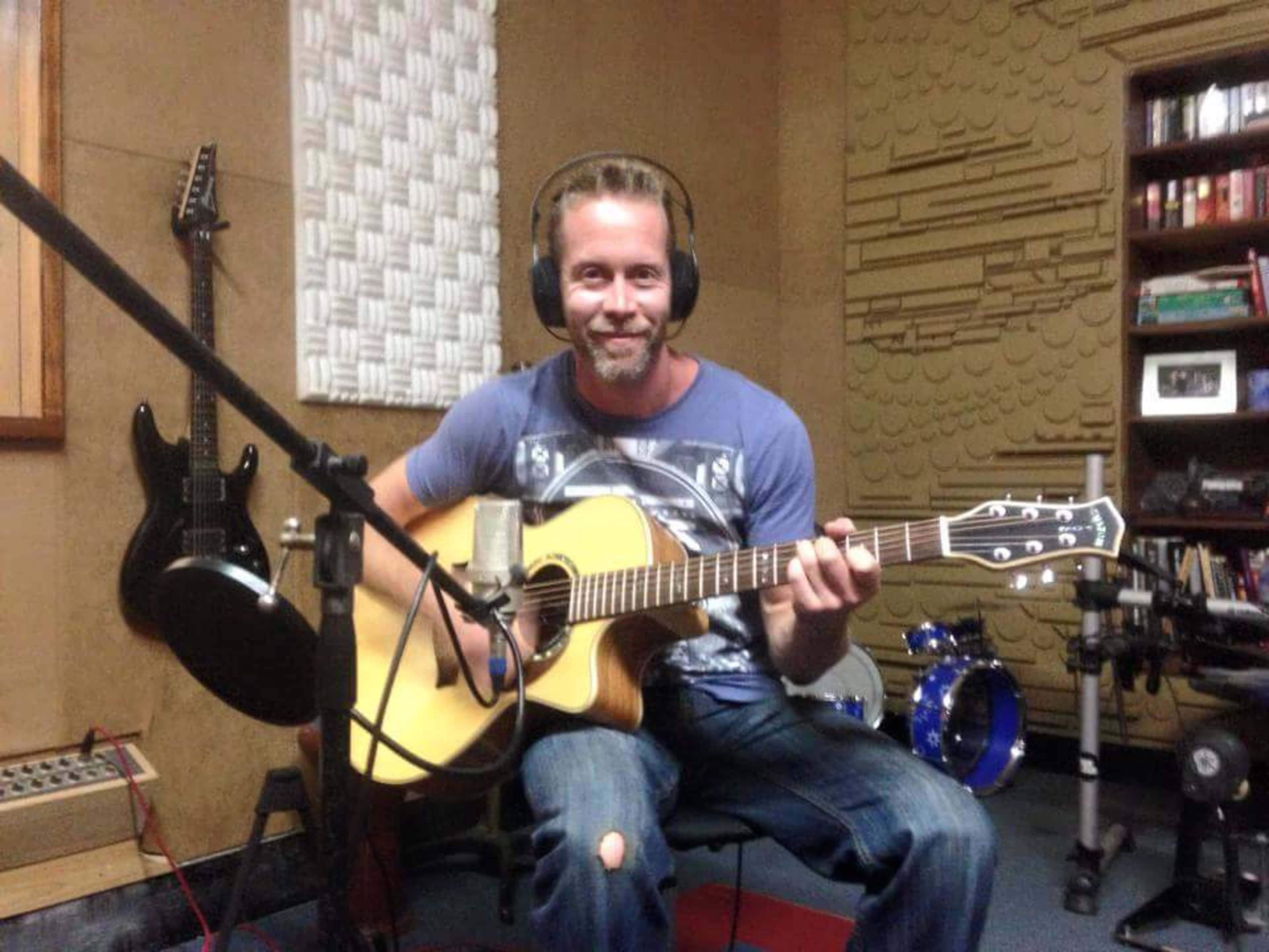 A man with headphones plays an acoustic guitar in a sound recording studio.