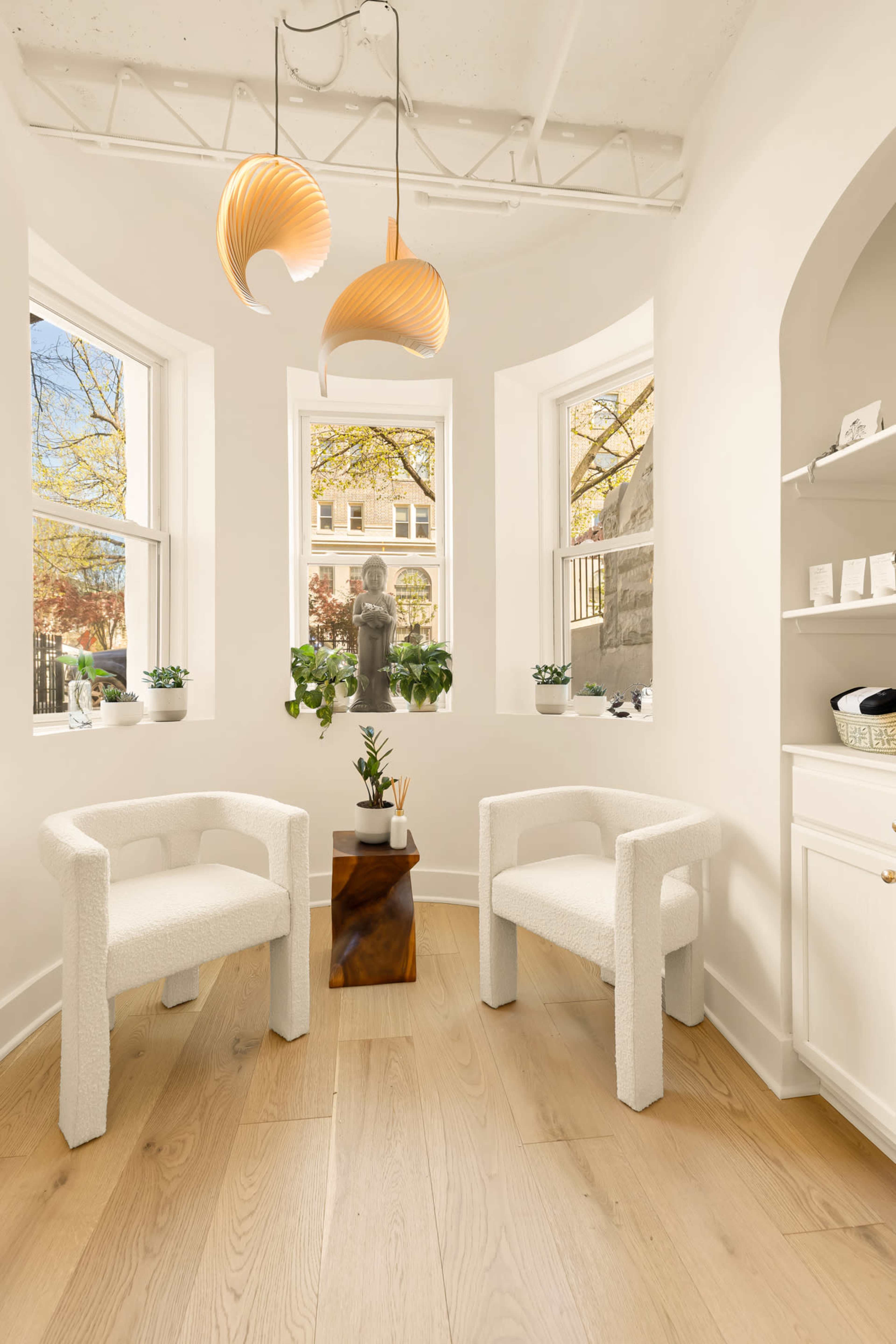 The image shows a bright, cozy seating area with two white chairs and a small wooden table, accented by a decorative light fixture and windows allowing natural light in.