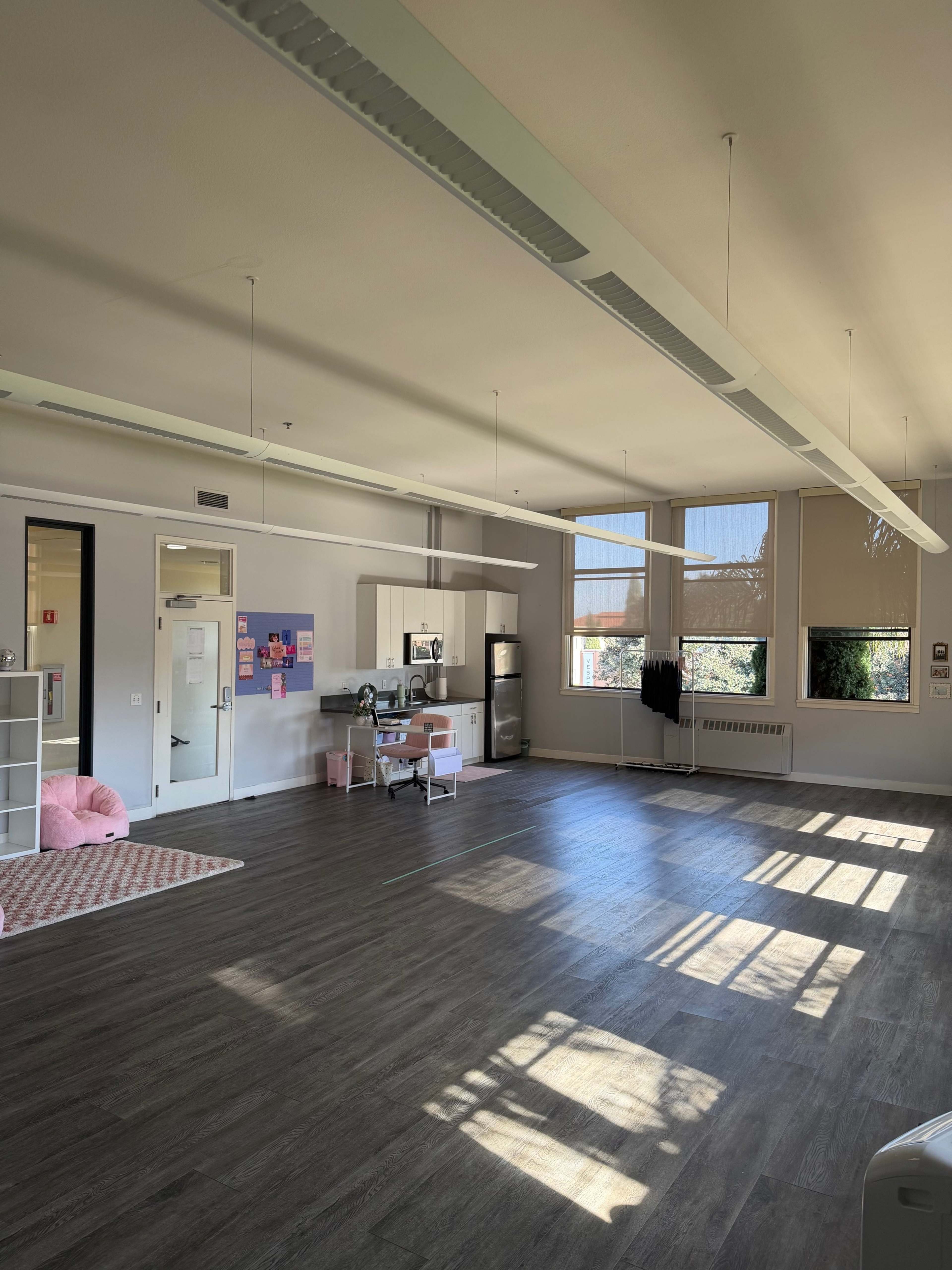 The image shows a spacious, brightly lit room with wooden flooring, featuring a kitchen area and large windows casting shadows on the floor.