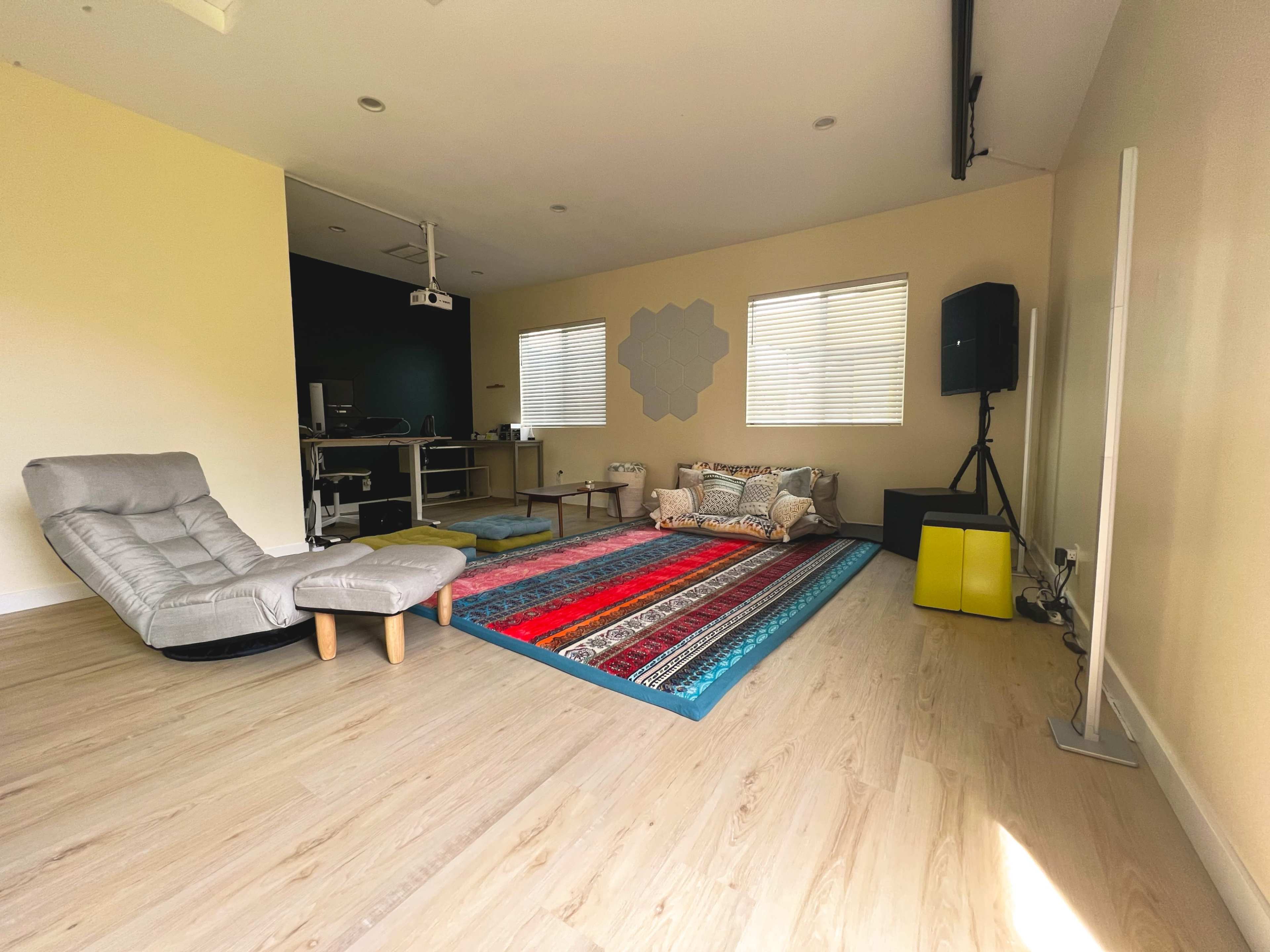 The image shows a spacious room with wooden flooring, featuring a large area rug, a seated lounge chair, and a small table, along with a projector setup and acoustic speakers in the background.
