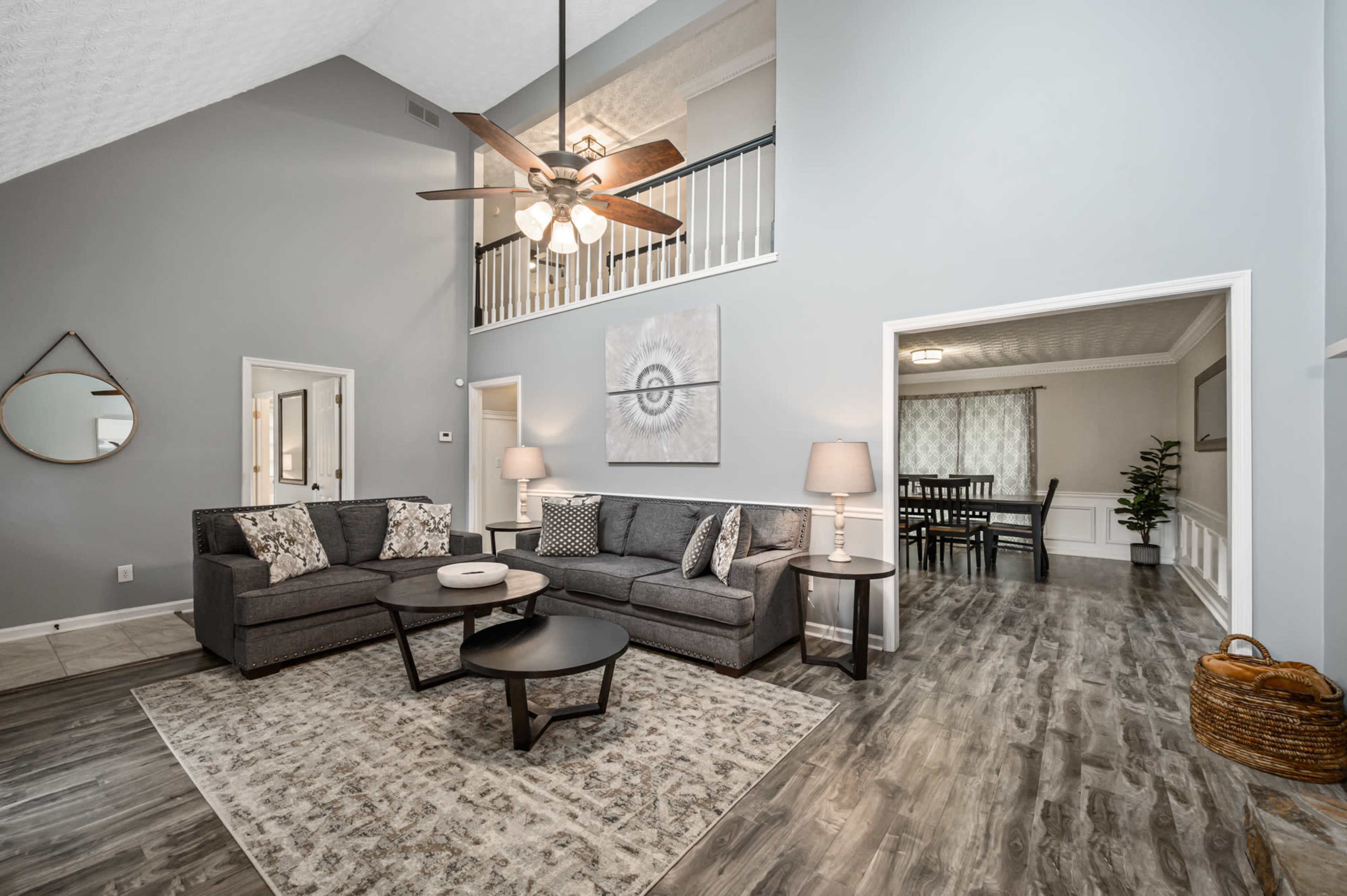 A modern living room with gray furniture, a circular coffee table, and a large rug, leading into a dining area with a visible table and chairs.