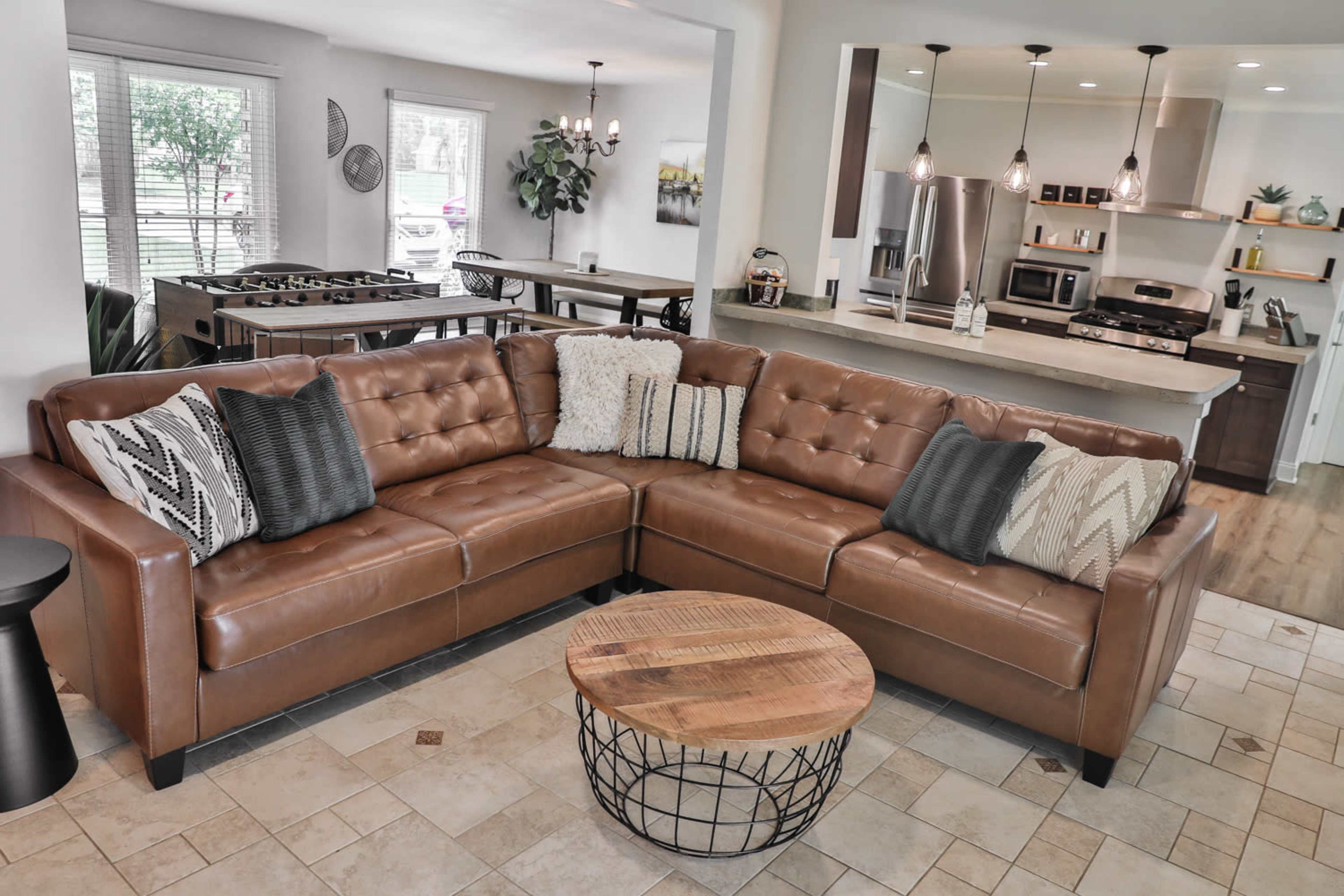 A modern living area featuring a brown leather sectional sofa with decorative pillows, a circular coffee table, and a view into a kitchen and dining space in the background.