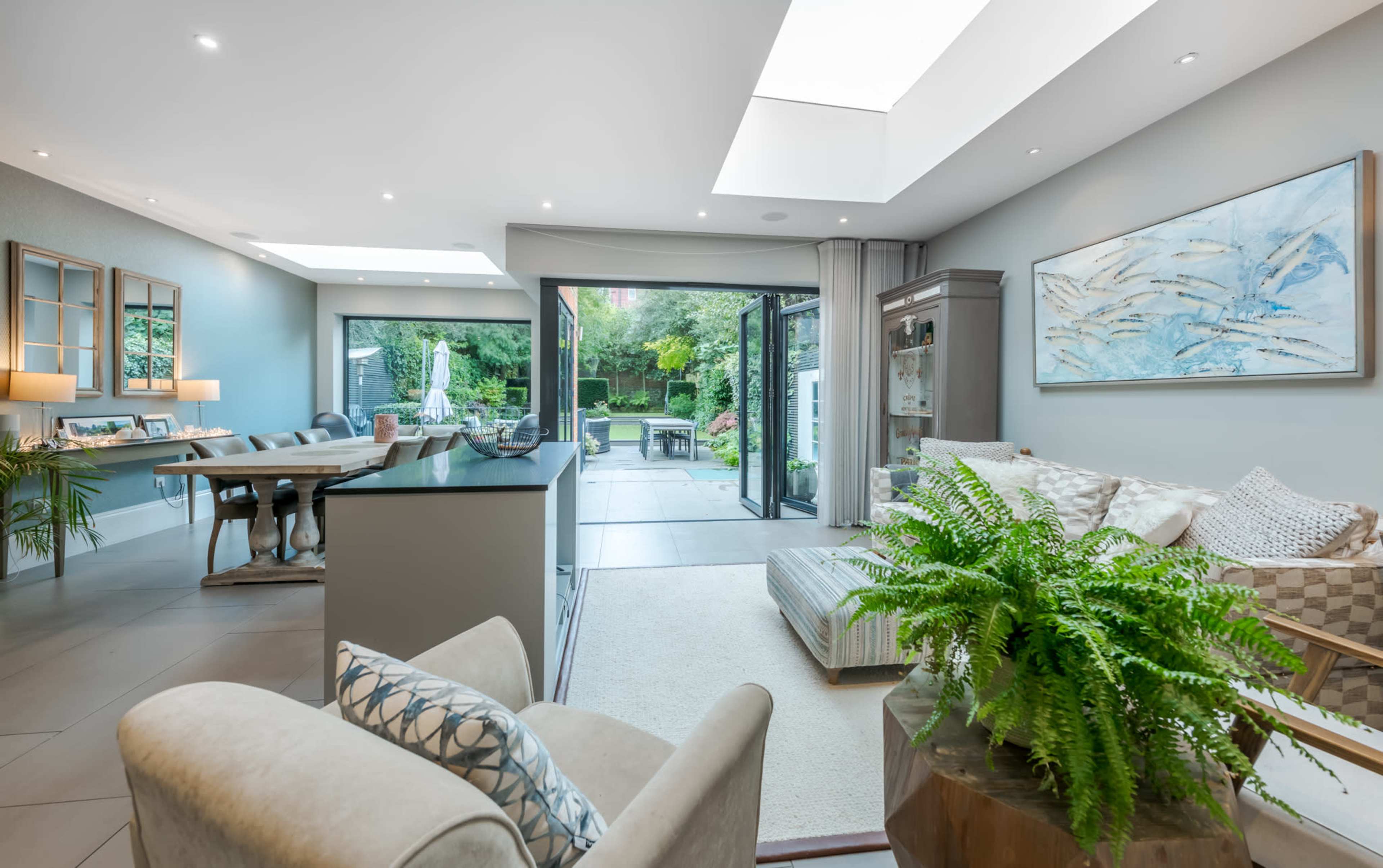 A modern living space features an open layout with a kitchen area, dining table, and a view of a garden through large sliding glass doors.