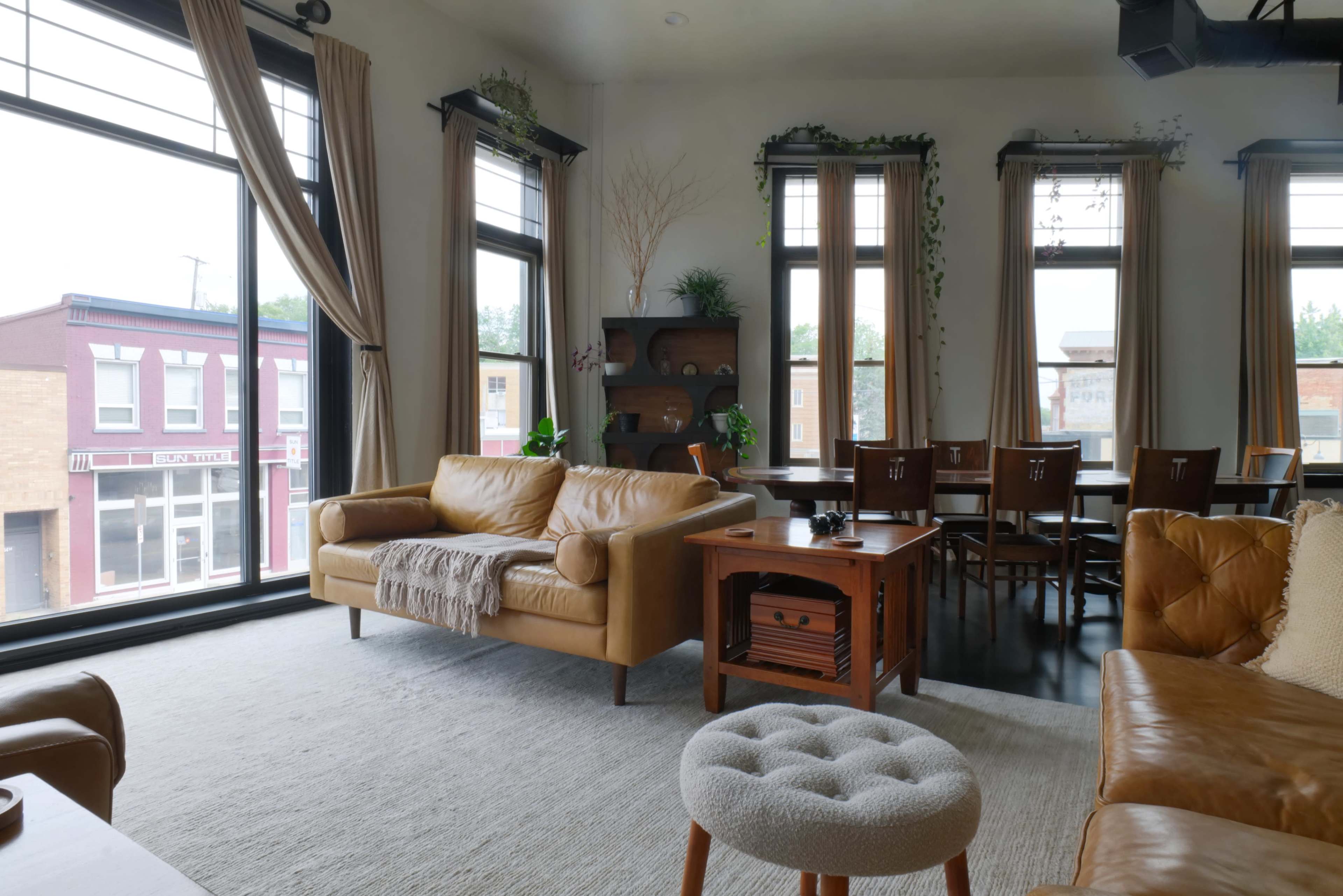 The image shows a cozy living room with large windows, a leather sofa, a wooden dining table, and indoor plants.