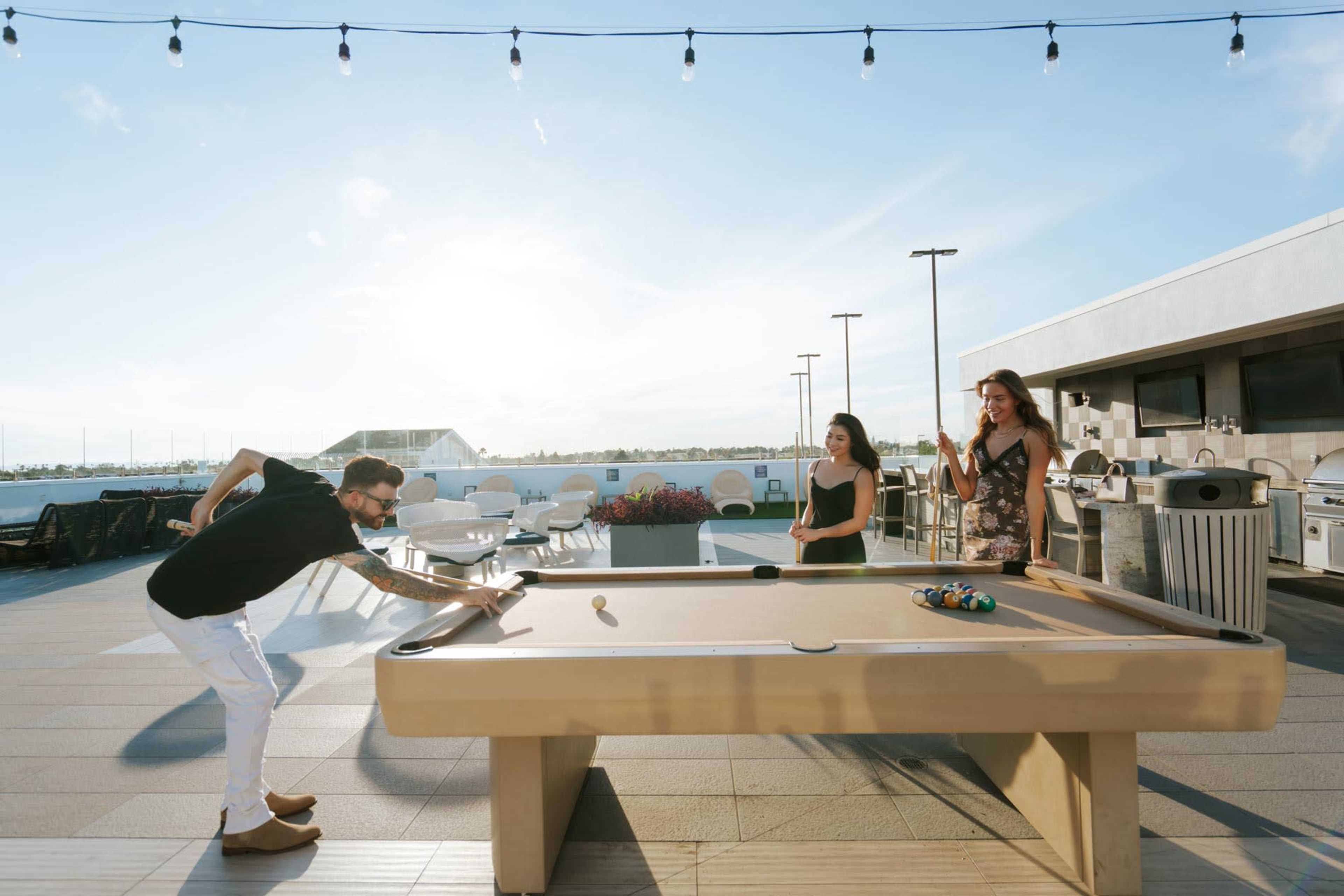 Three people are outside on a rooftop playing pool with a view of the sky and surrounding area.