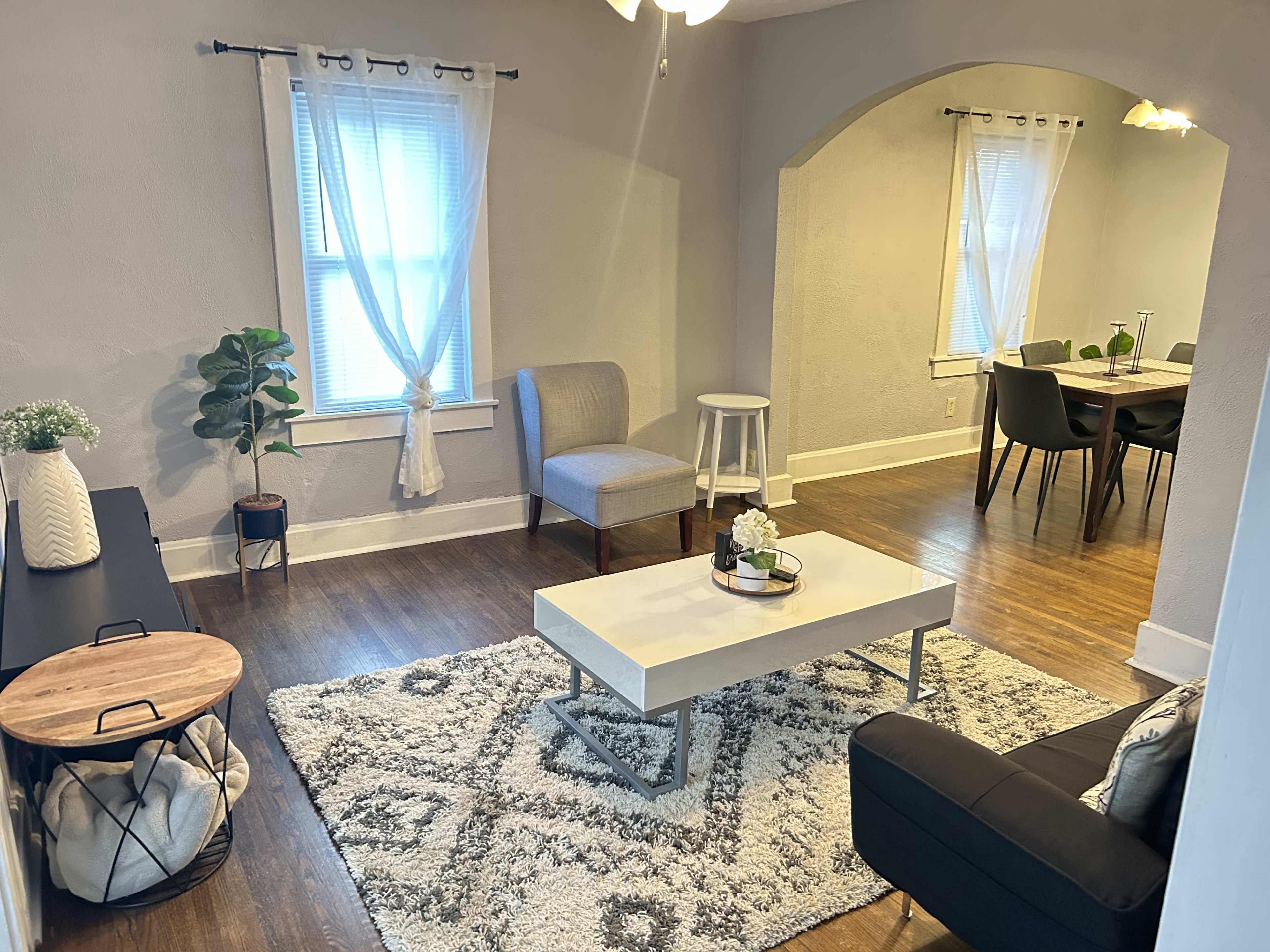A cozy living room with a gray color scheme, featuring a coffee table, chairs, and a plant, connected to a dining area in the background.