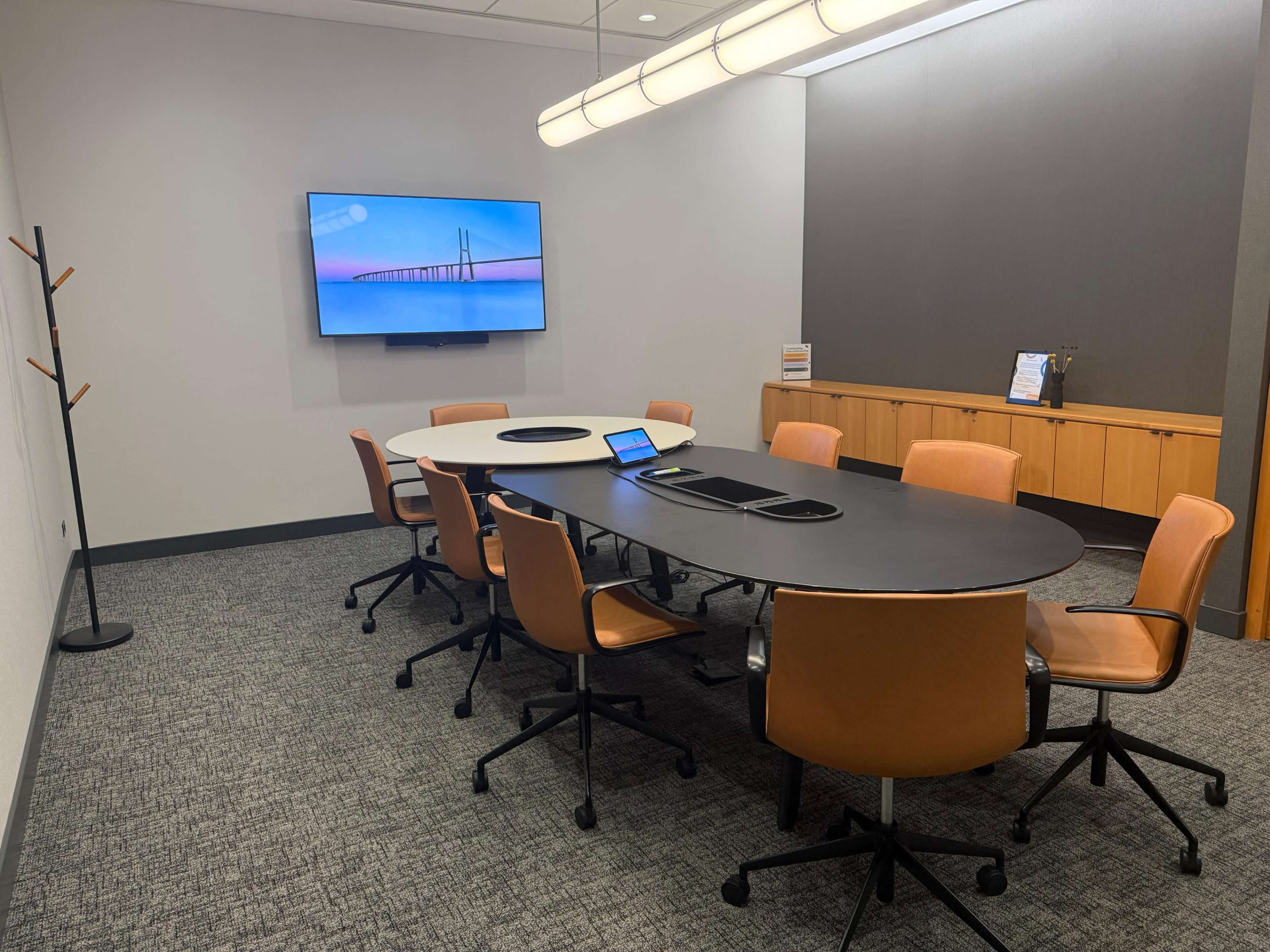 The image shows a modern conference room featuring a large oval table surrounded by eight brown leather chairs, a wall-mounted screen displaying an abstract image, and a wooden cabinet along one wall.