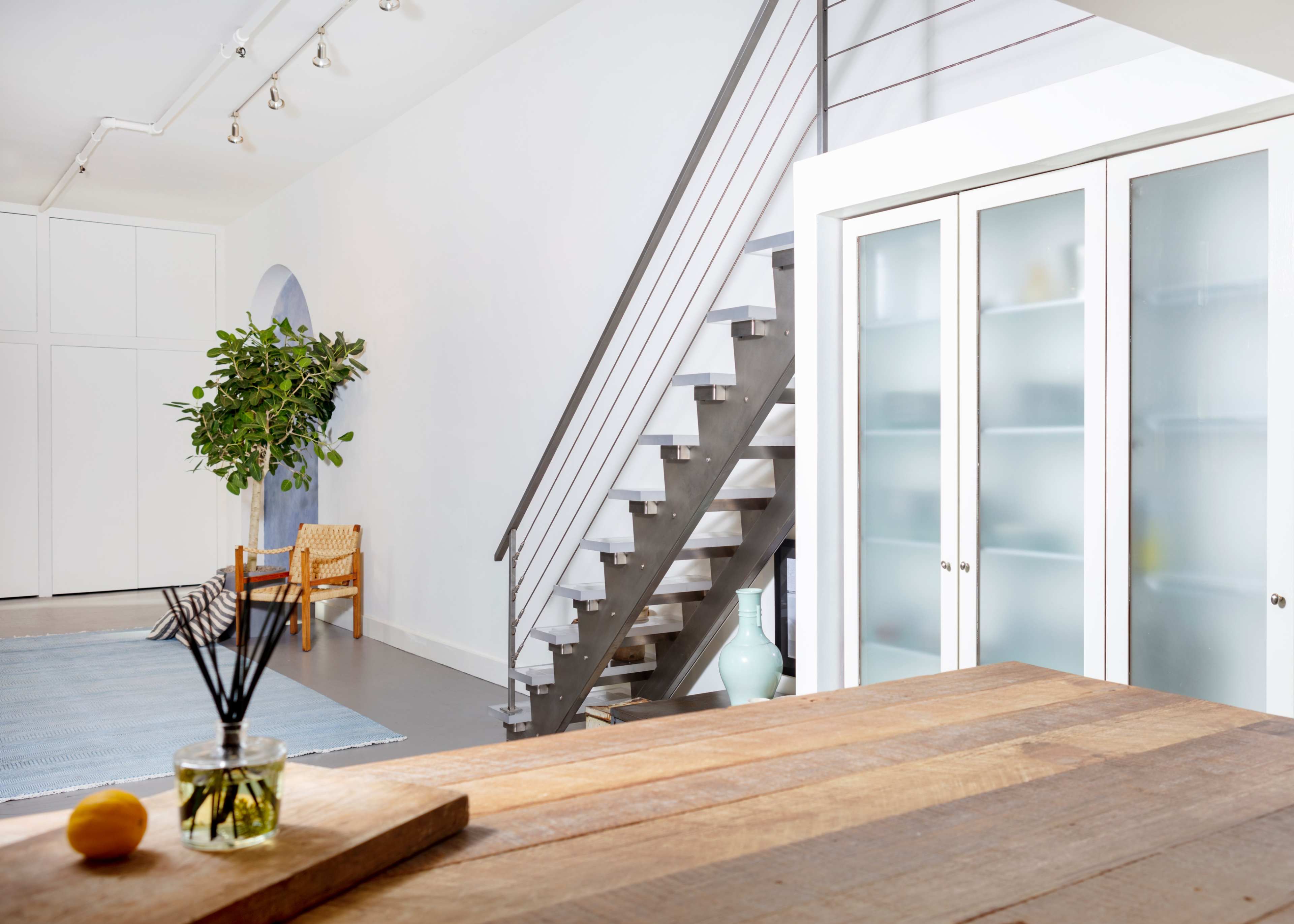 A modern interior space features a staircase, a glass-door cabinet, a plant, and a wooden table in a well-lit room.