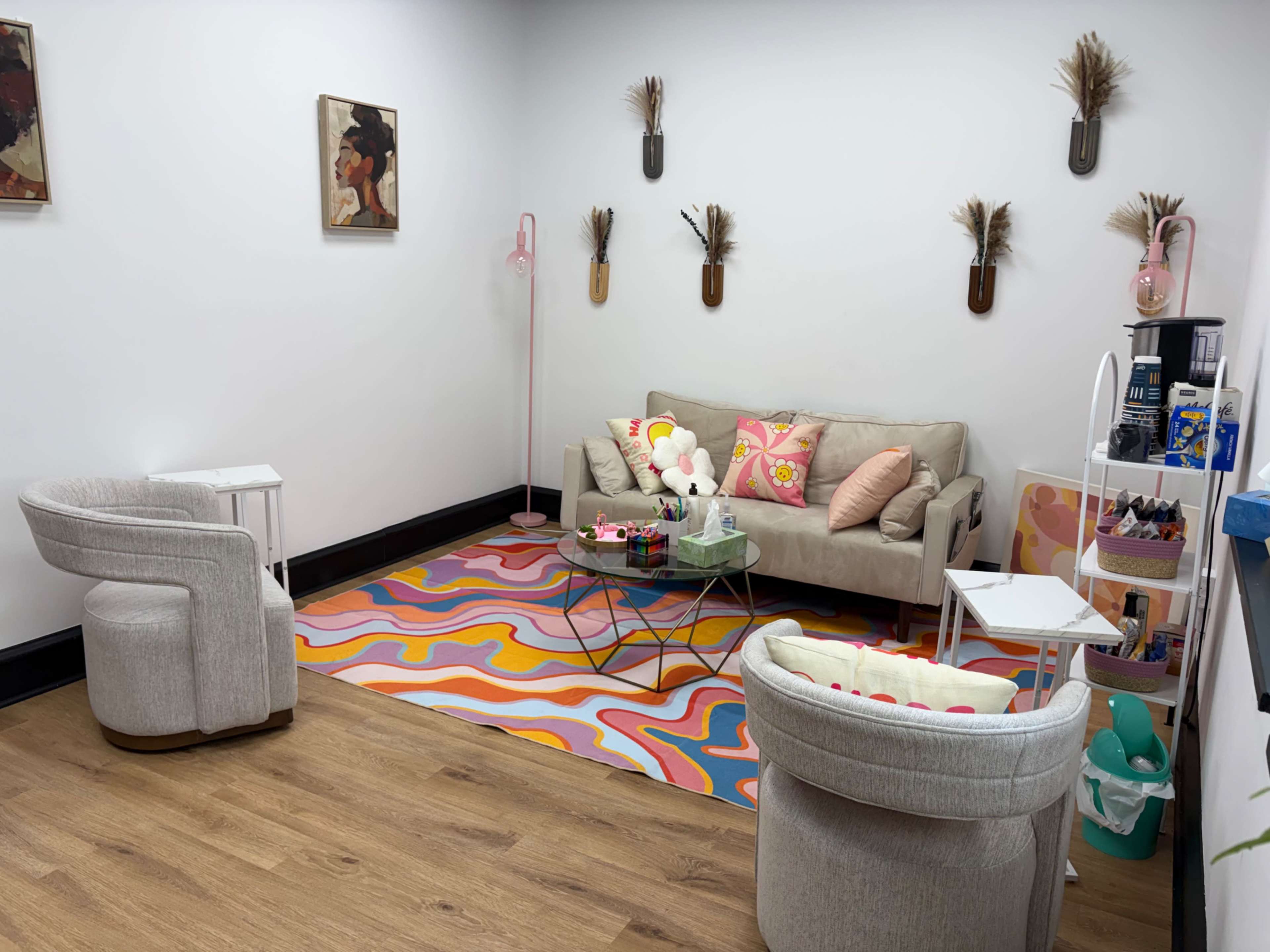 The image shows a brightly decorated waiting area featuring a beige sofa and two light-colored chairs on a colorful patterned rug, with decorative elements on the walls and a side table displaying various items.