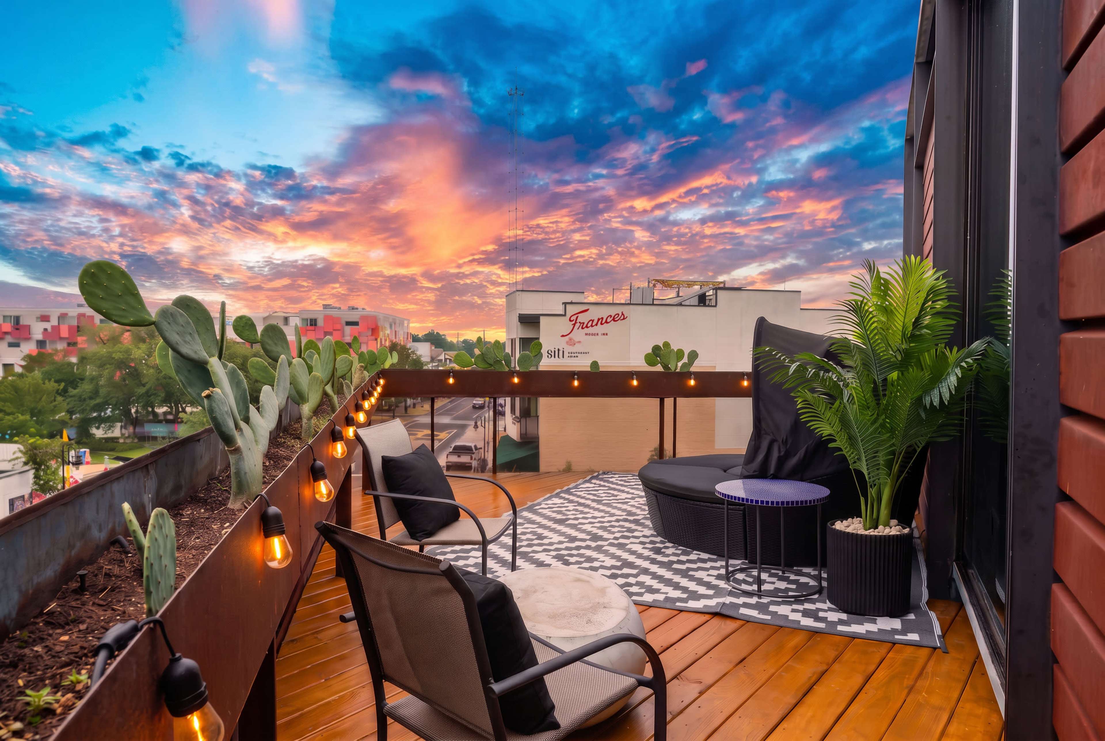 The image shows a wooden balcony with a black and white patterned rug, two chairs, a round table, potted plants, and string lights, set against a vibrant sunset sky.