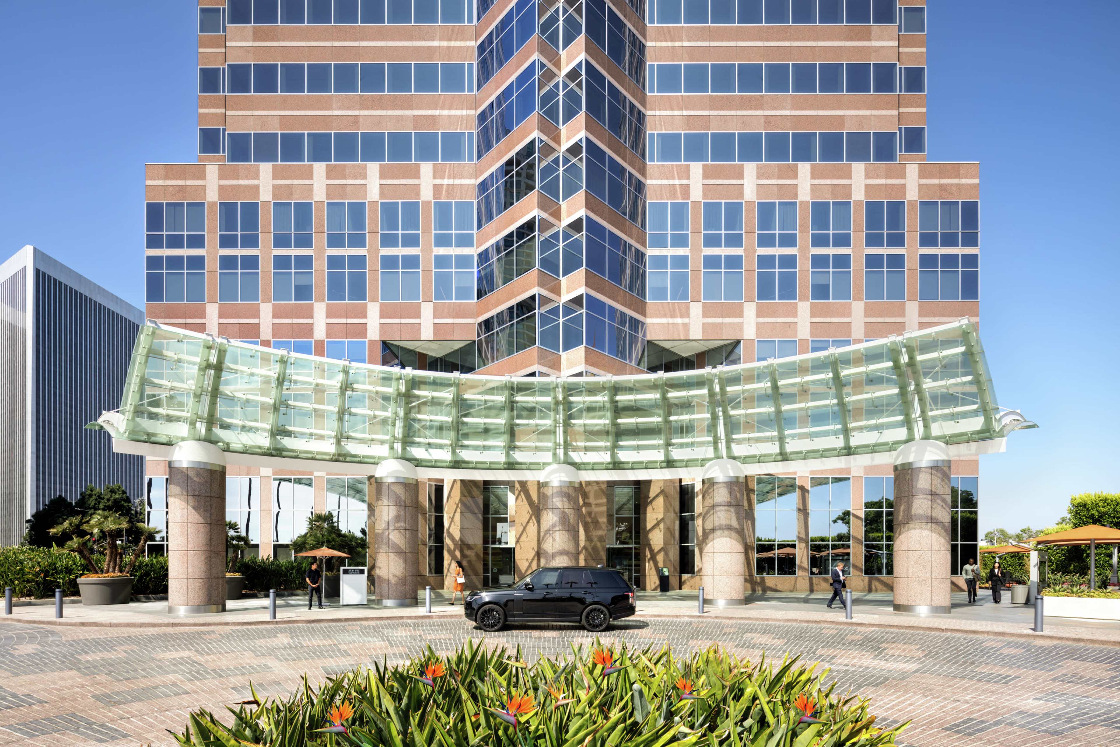A modern office building with a glass canopy entrance and landscaped greenery in the foreground.