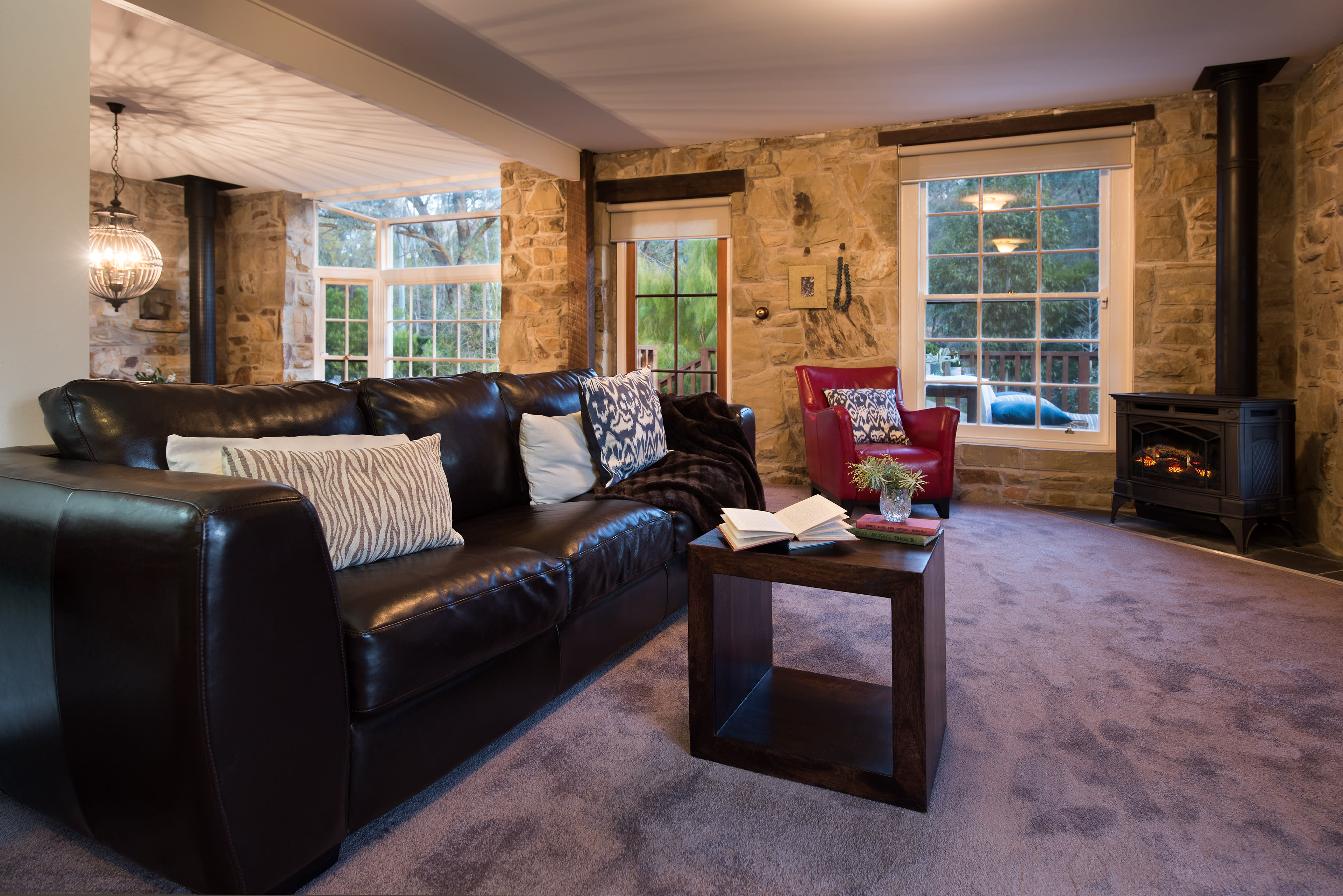 A cozy living room features a dark leather sofa, a red armchair, and a wooden table with an open book, surrounded by stone walls and large windows.